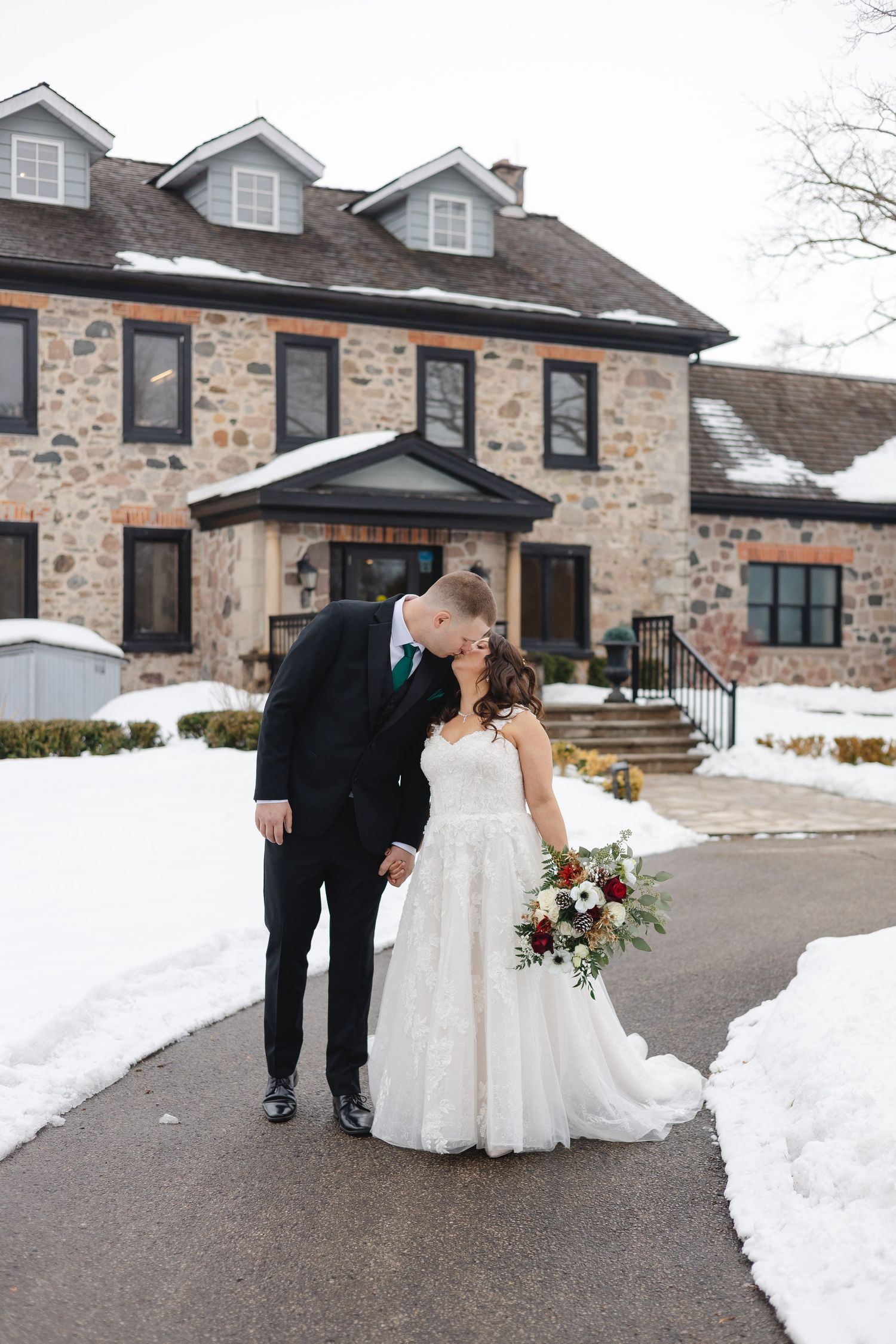 Bride and groom sharing a kiss in front of a stone building surrounded by snow, bride holding a red and white bouquet.