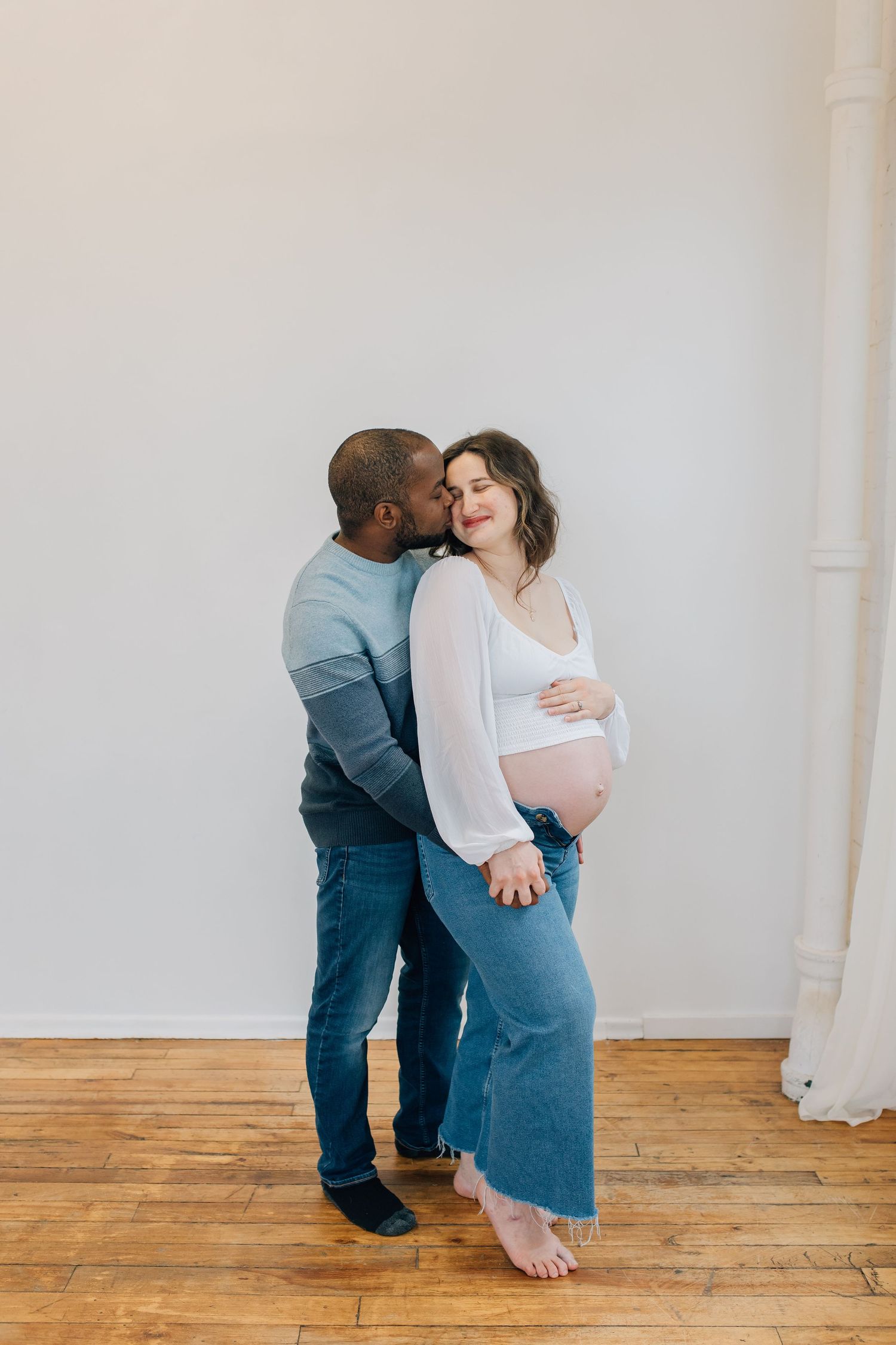 Interracial couple sharing kiss as man embraces pregnant woman holding her baby bump indoors.