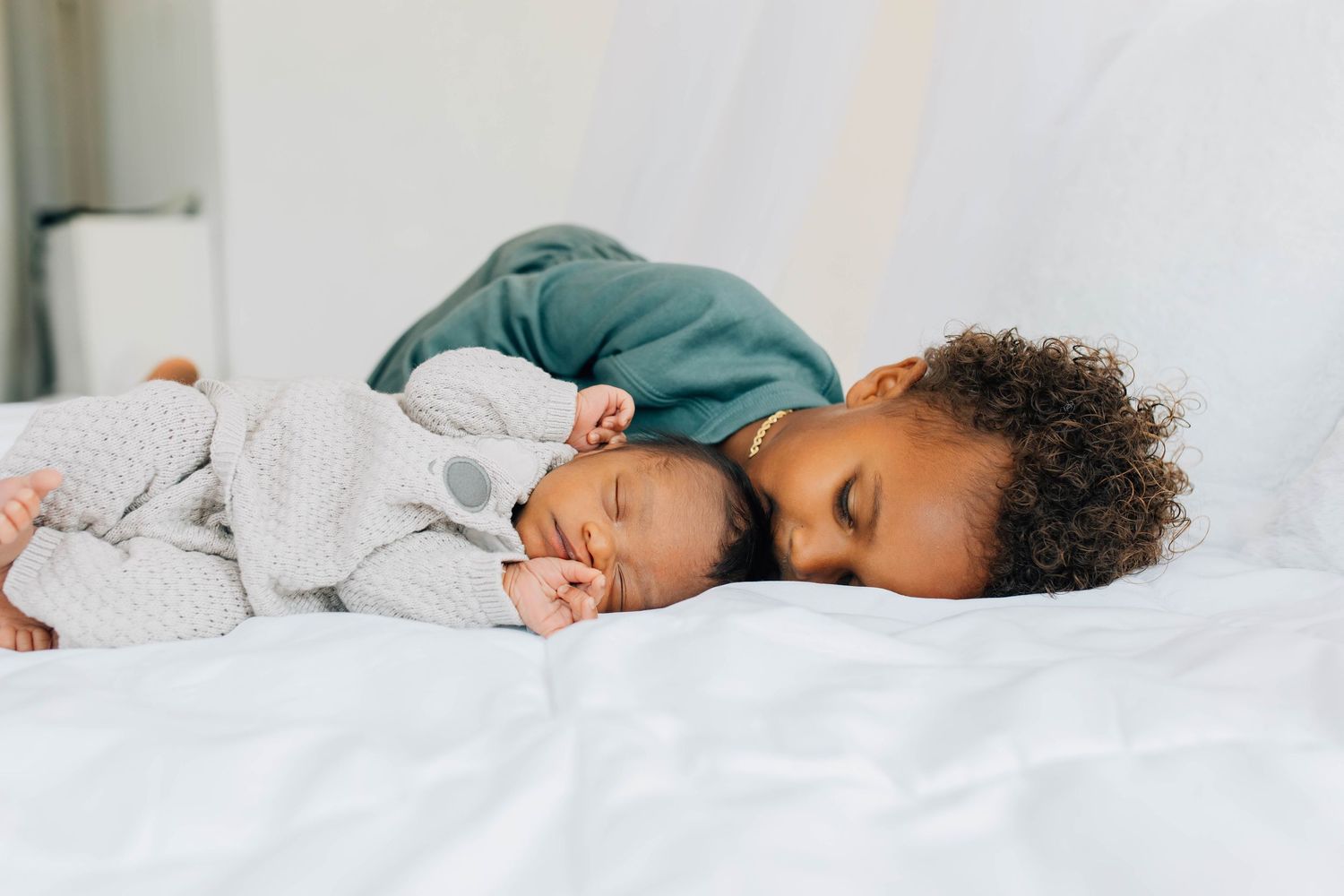 Sweet toddler in green outfit gently resting head on sleeping newborn sibling on white bed.