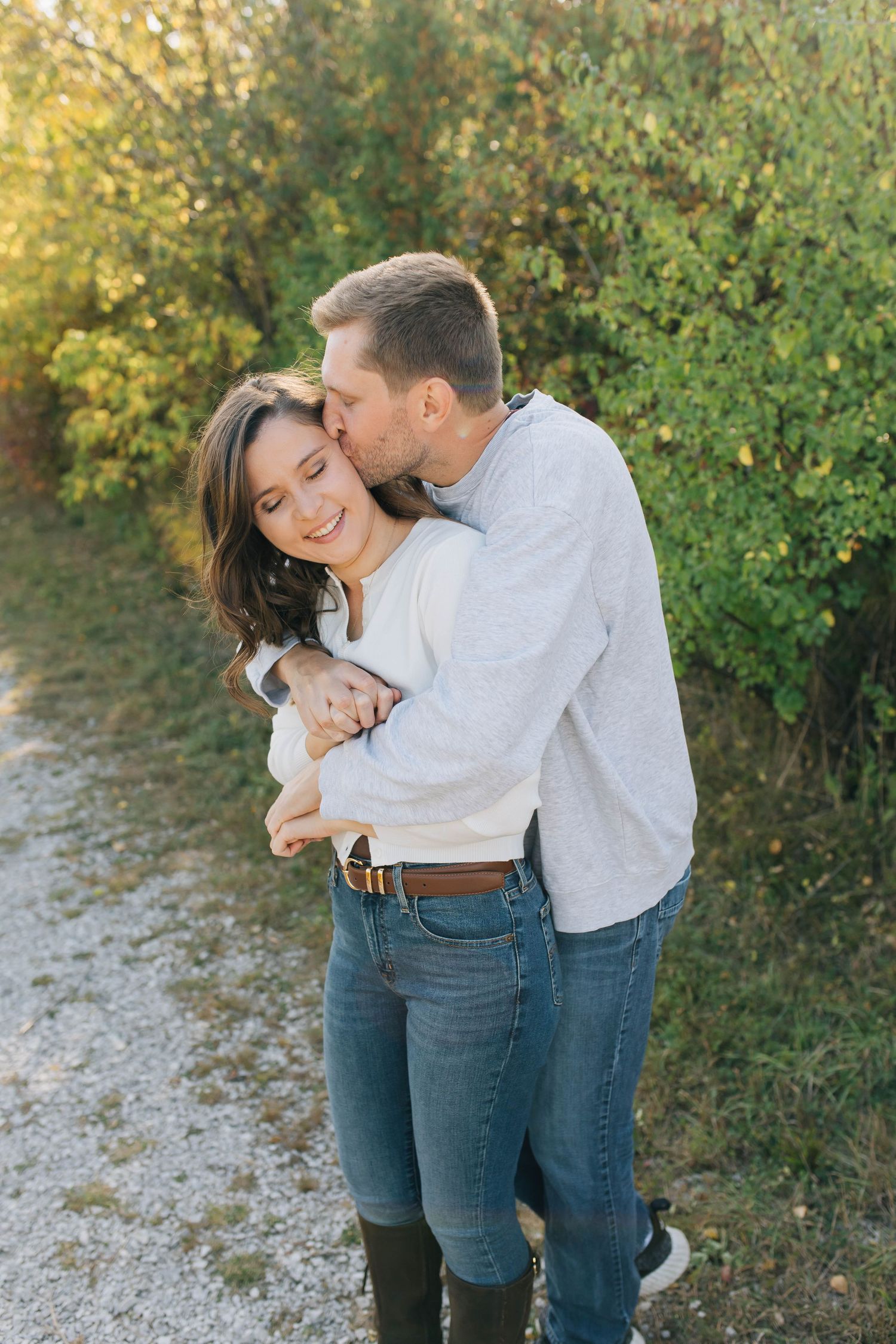 Man kissing woman on forehead while embracing on a sunlit gravel path with greenery.