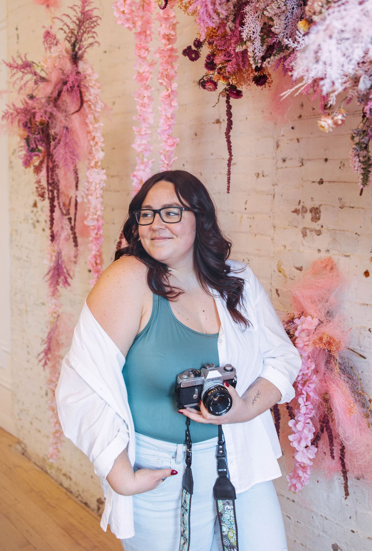 Woodstock Ontario wedding photographer smiling and holding a film camera in front of pink floral wall decor.