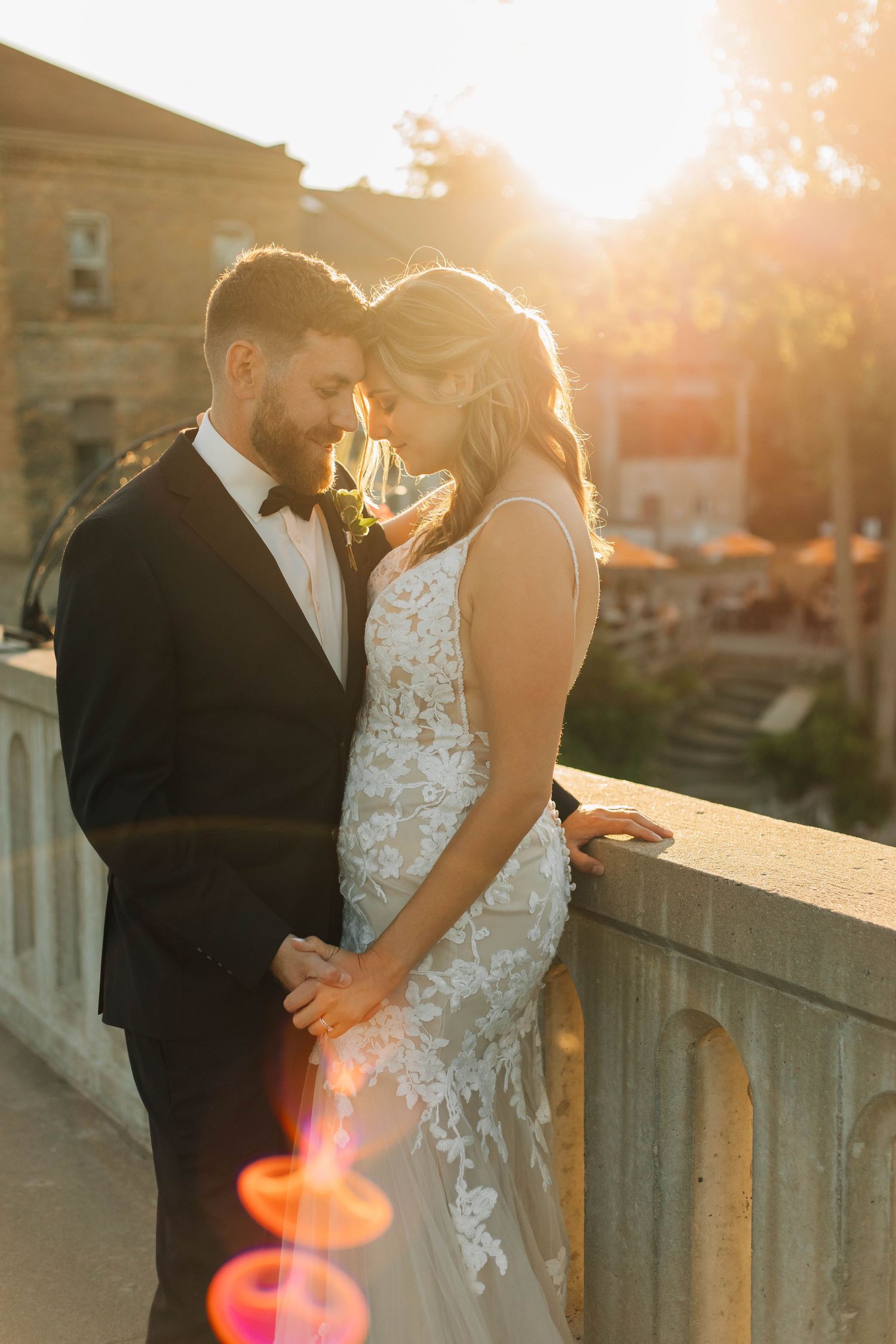Bride and groom share a romantic golden hour moment on a stone bridge at sunset.