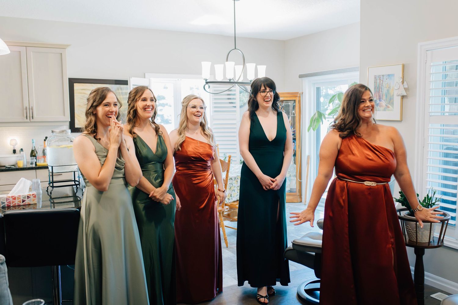 Five bridesmaids in colorful satin gowns laughing together in a bright kitchen before a wedding ceremony.