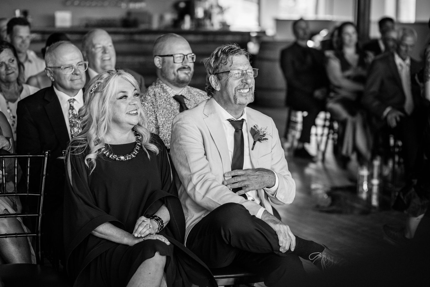 Wedding guests laughing and smiling while seated at indoor ceremony in black and white.