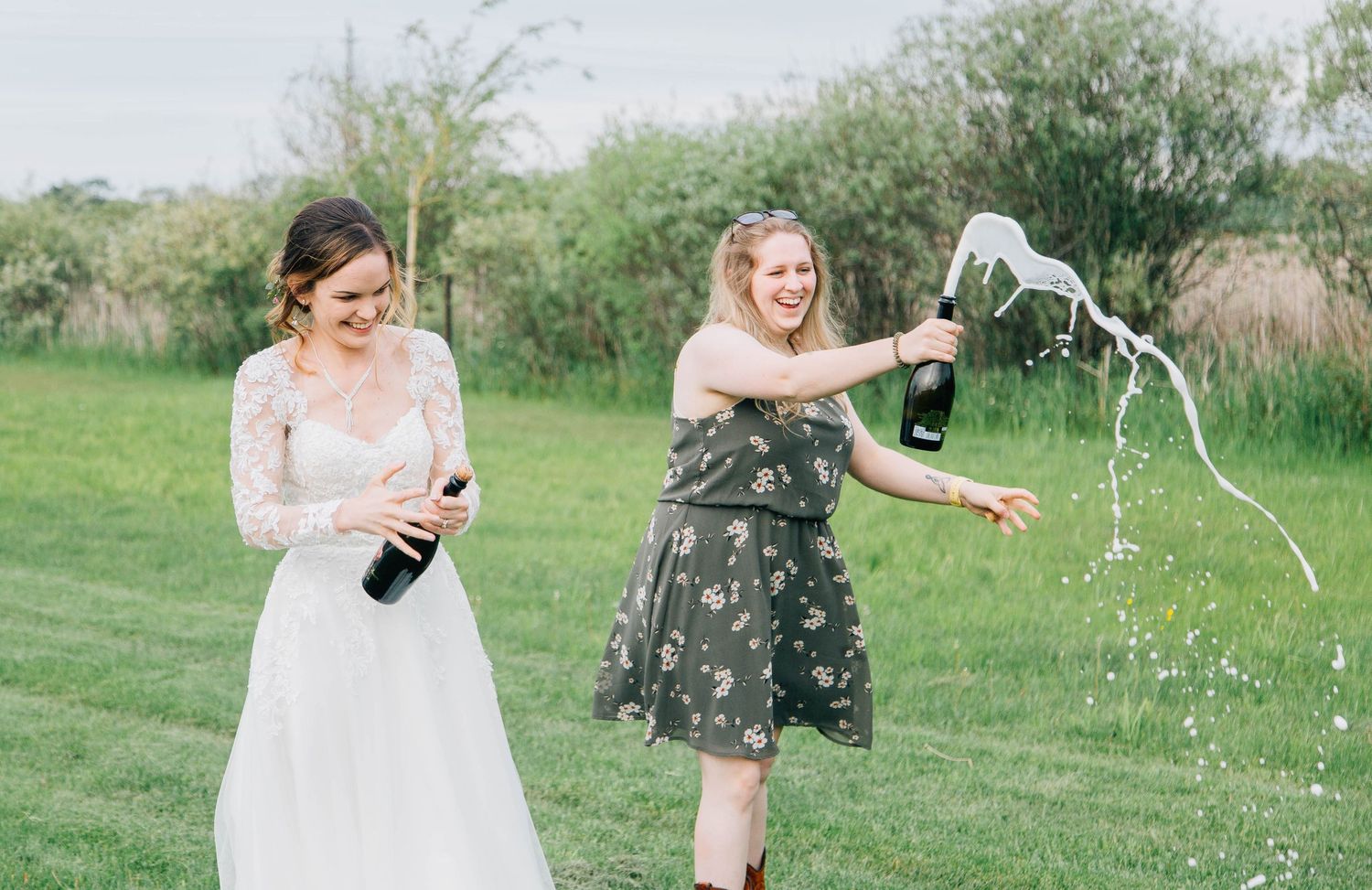 Bride and bridesmaid joyfully popping champagne bottles outdoors at a wedding celebration.