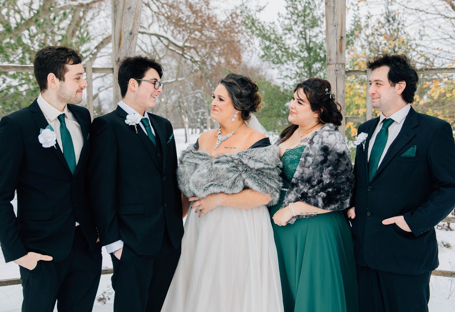Bride in white gown with fur wrap laughs with wedding party guests in emerald green and black.