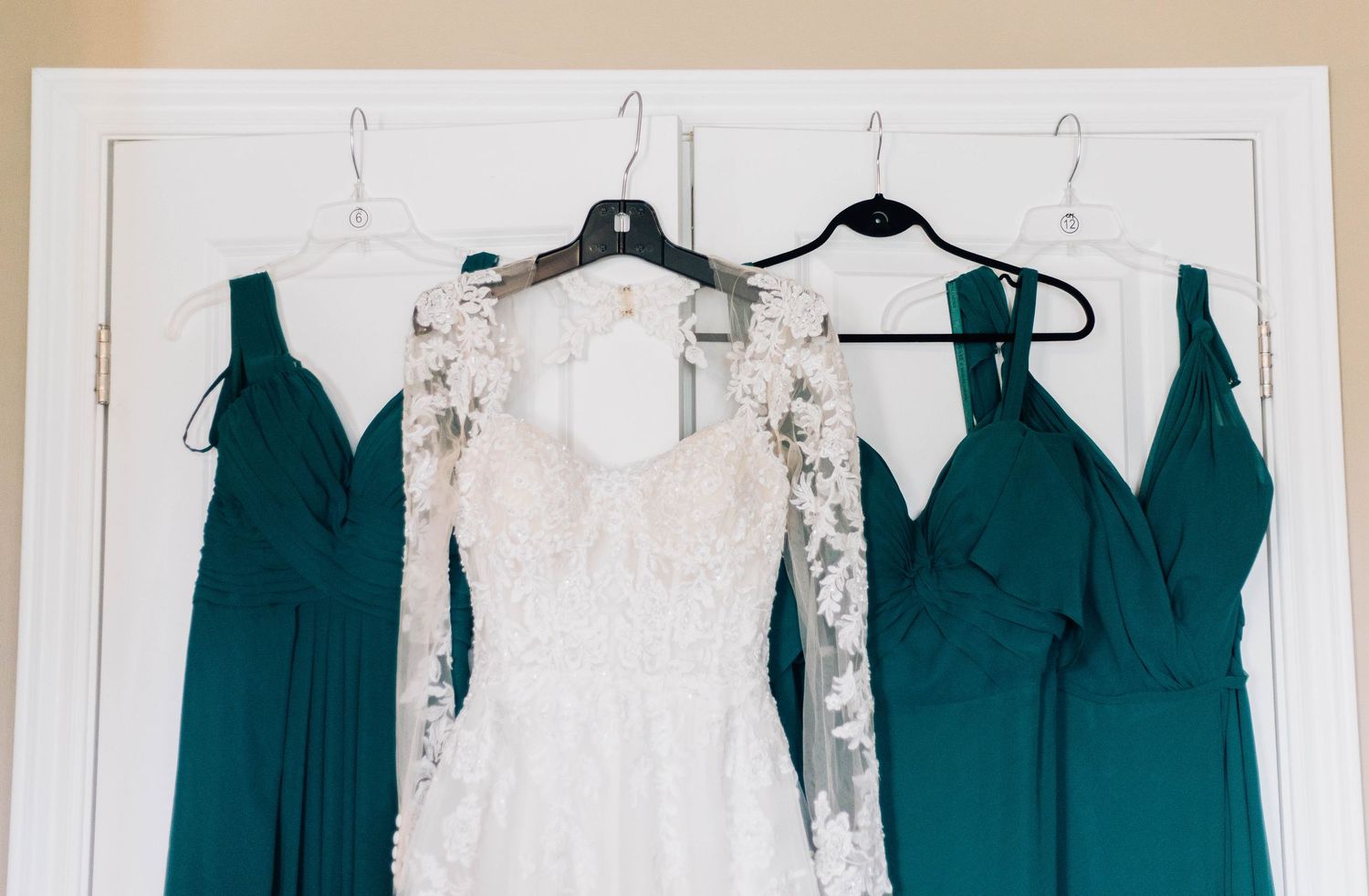 Lace wedding dress flanked by two teal bridesmaid dresses hanging on white doors.