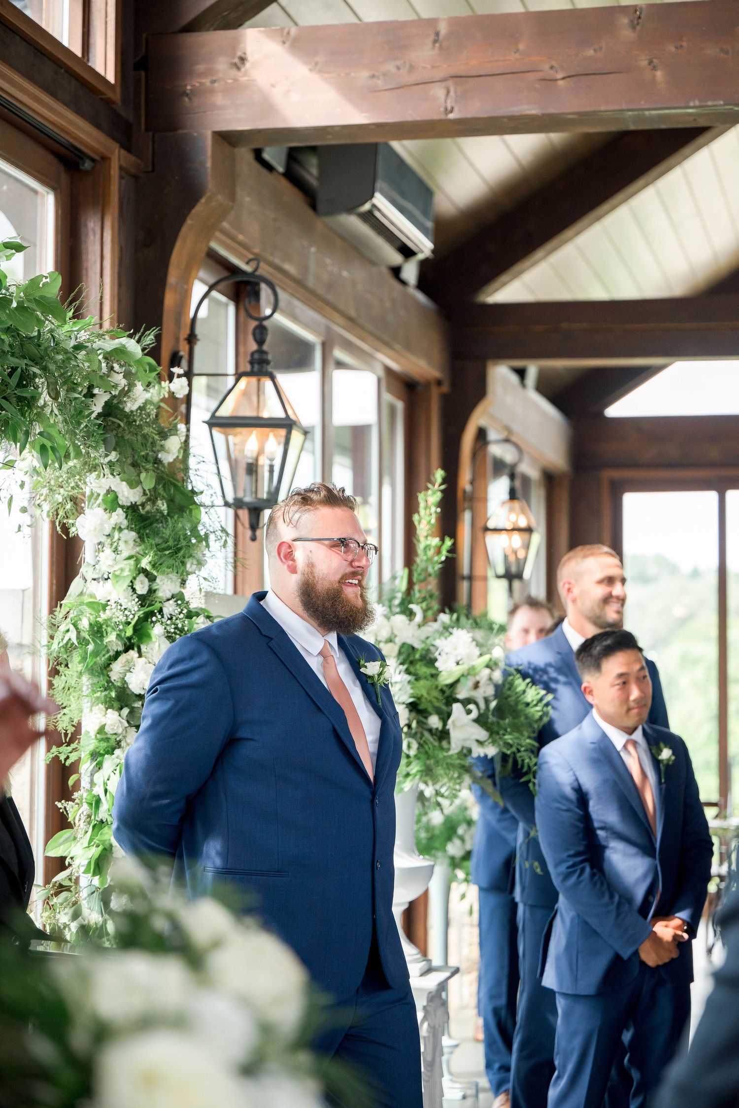Groom in navy suit watches bride walk down aisle, groomsmen standing beside floral ceremony arch.