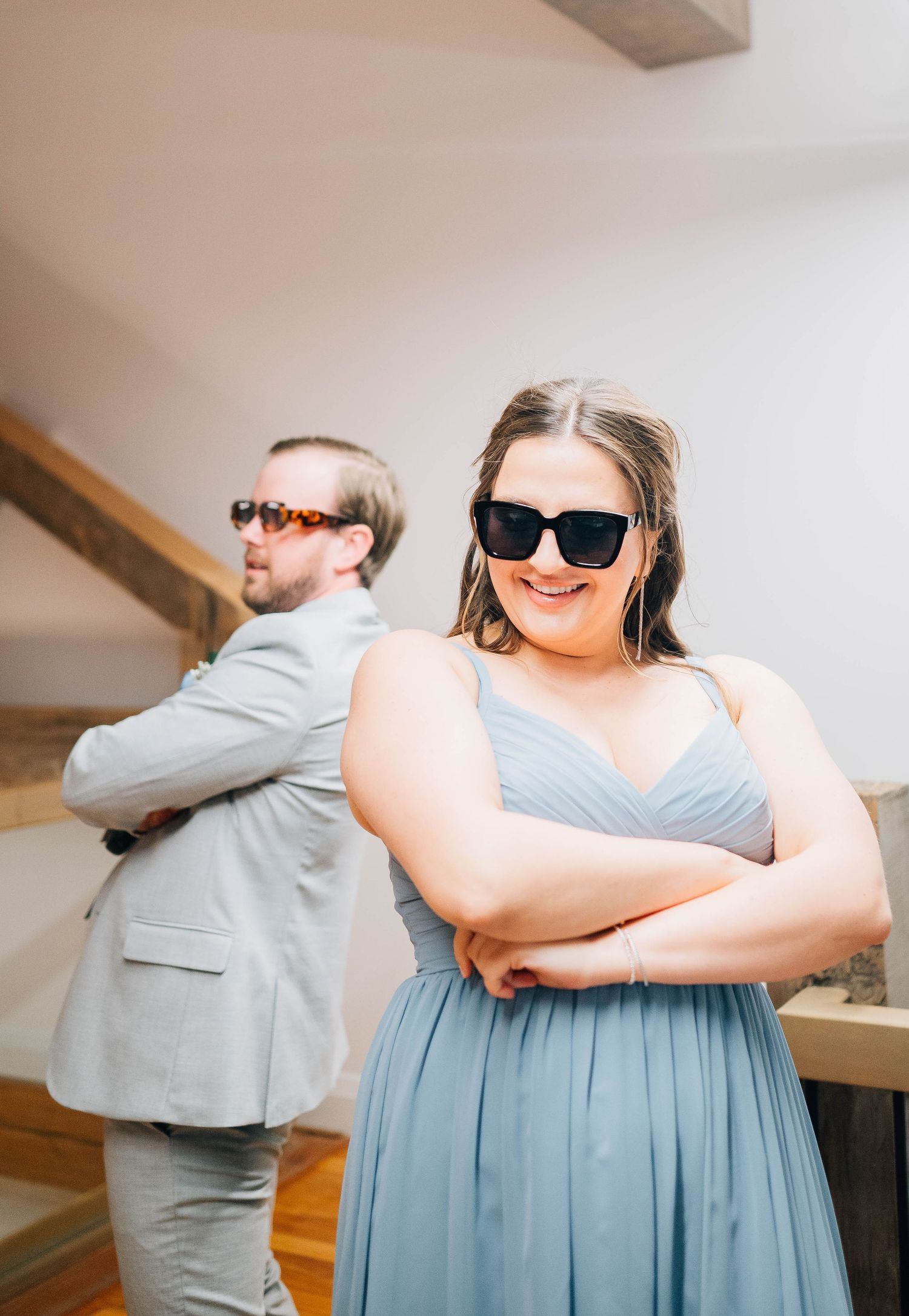 Bridesmaid and groomsman in sunglasses posing back-to-back with arms crossed at wedding celebration.