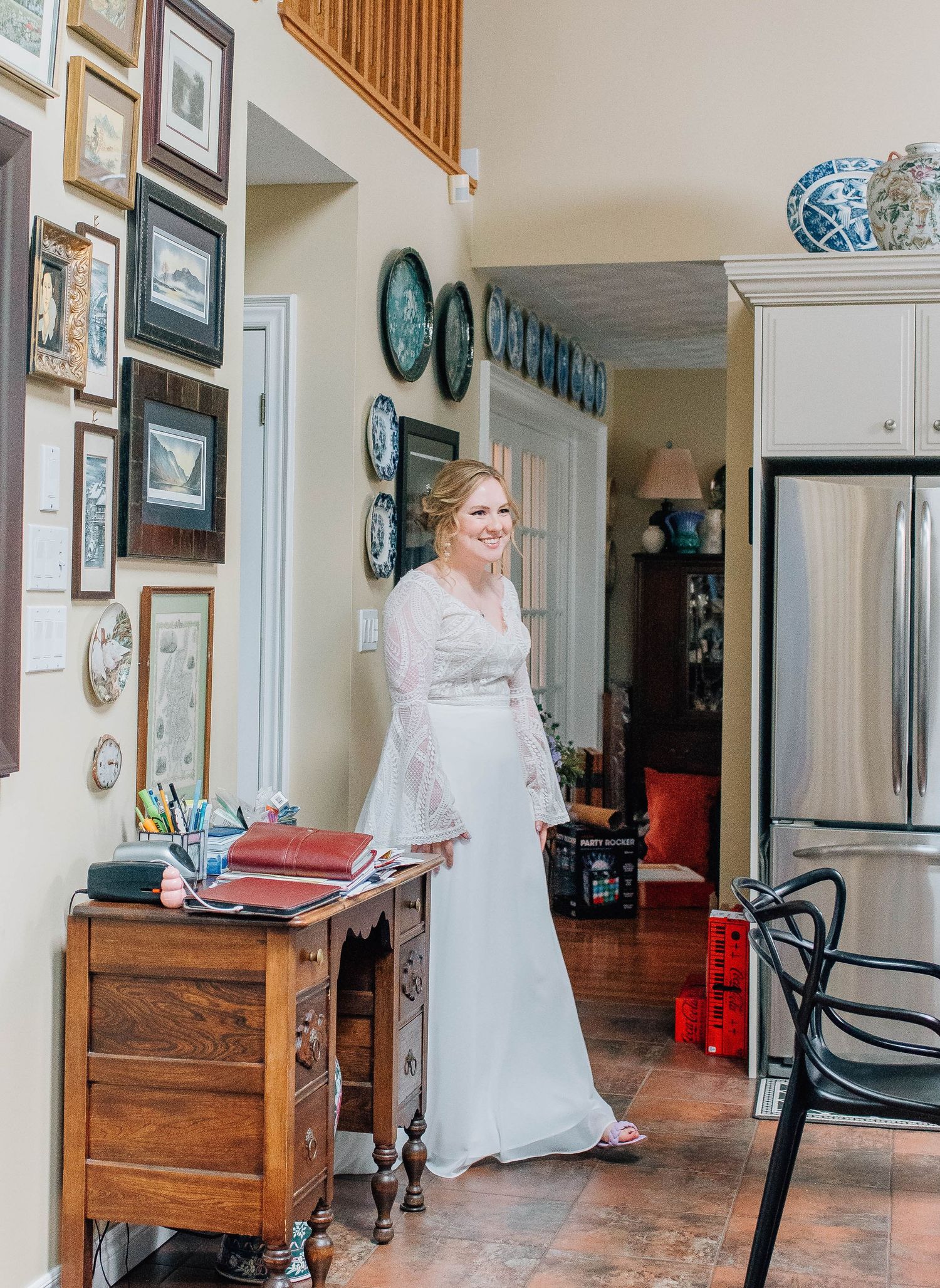 Bride in elegant white lace gown stands in rustic home hallway with gallery wall.
