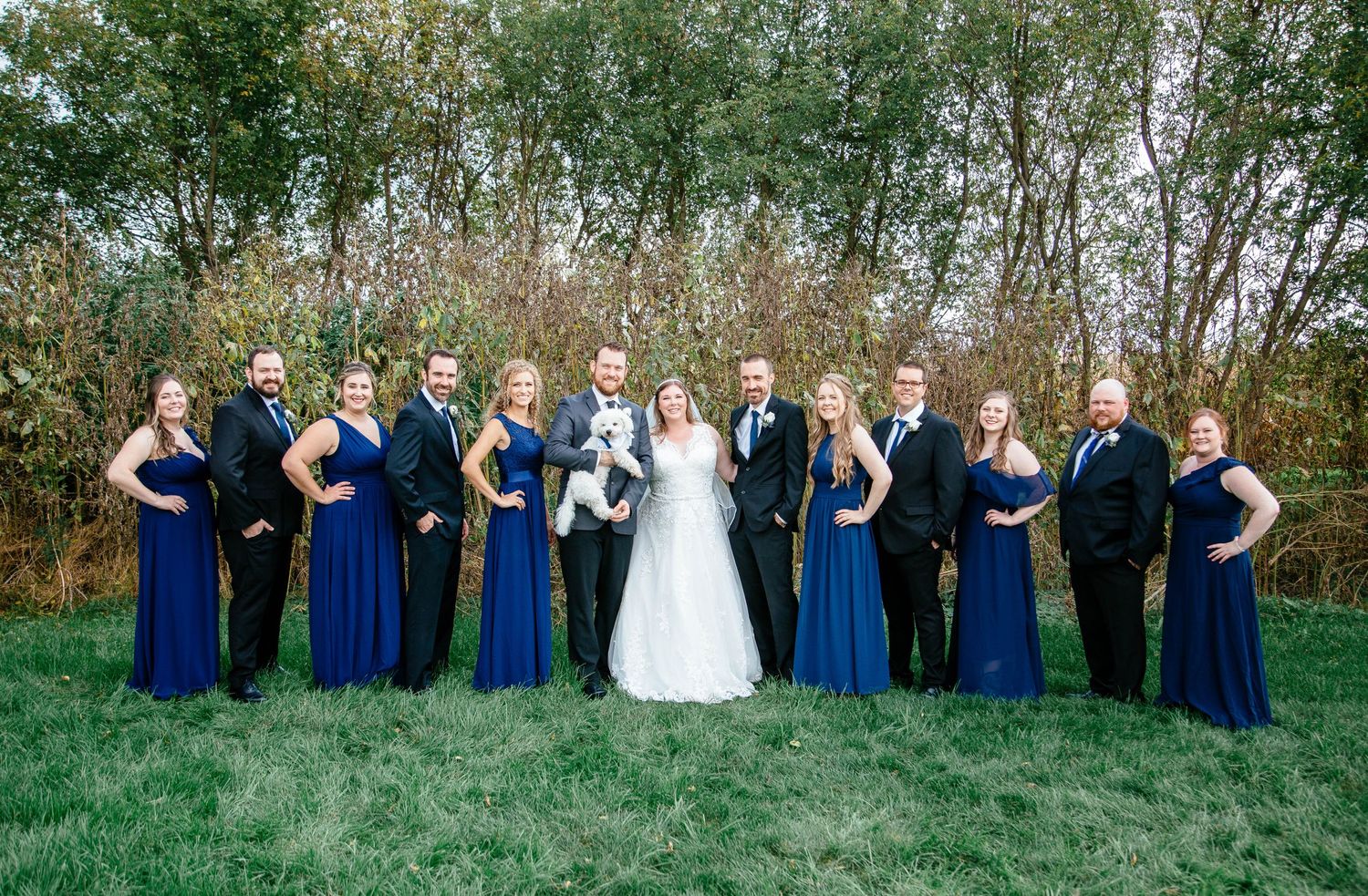 Wedding party in navy blue dresses and suits posing with bride and groom on green lawn.