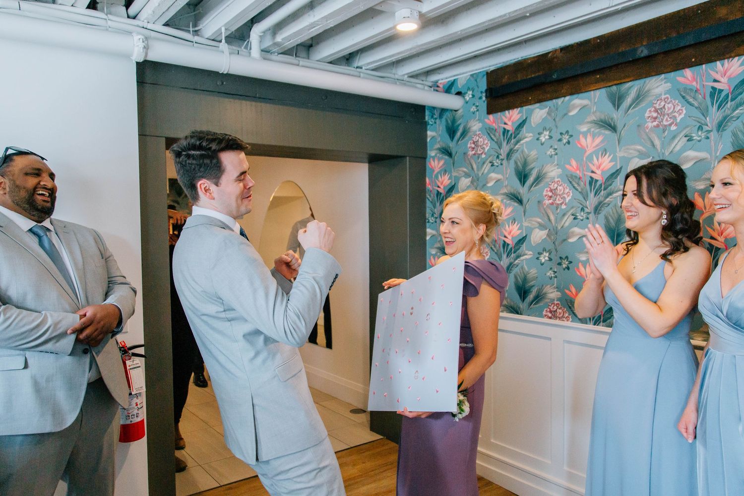 Groom pumps fist in celebration while bridesmaid holds poster and guests applaud at indoor wedding venue.