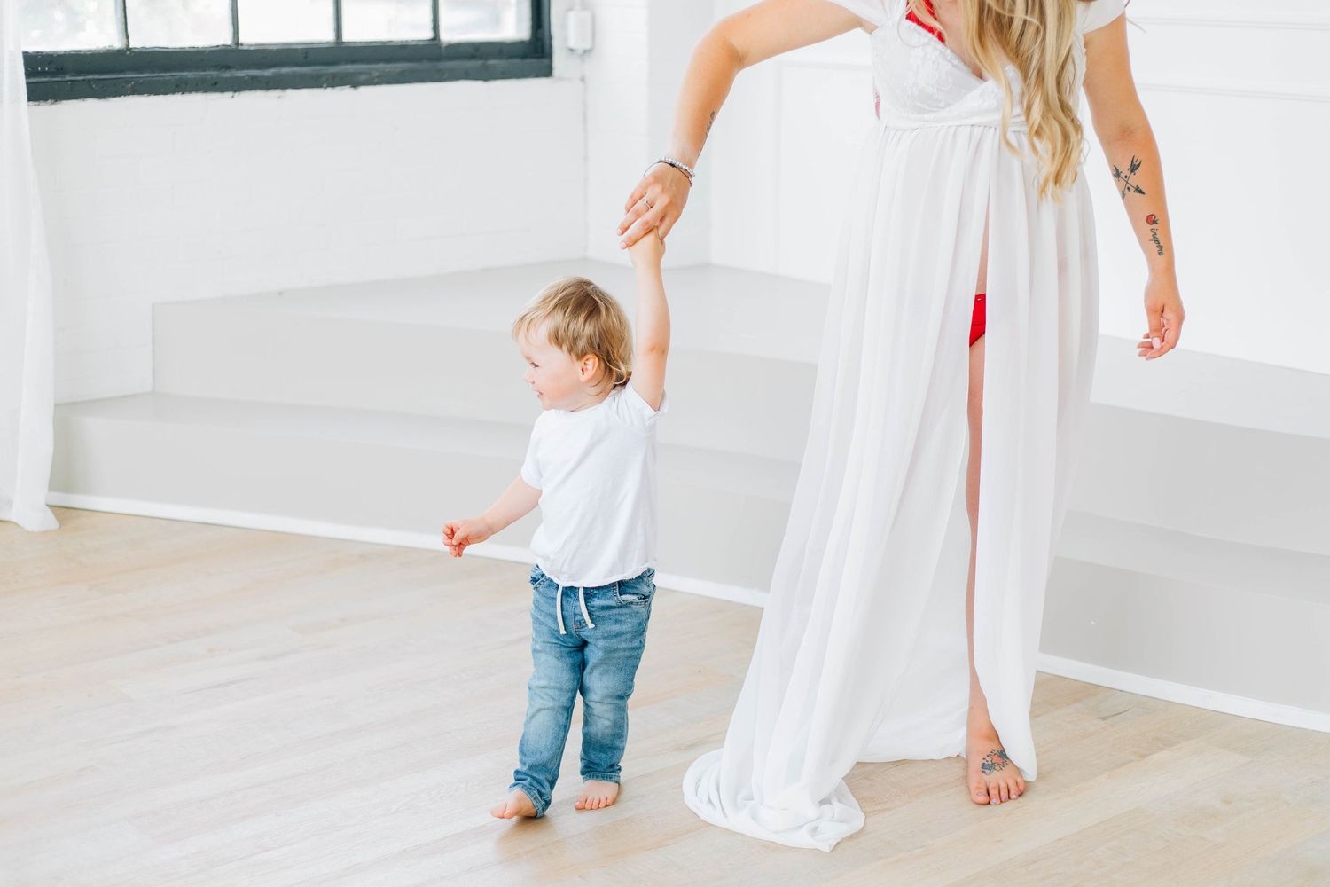 Pregnant mother in white dress holding toddler's hand on light wood floor.