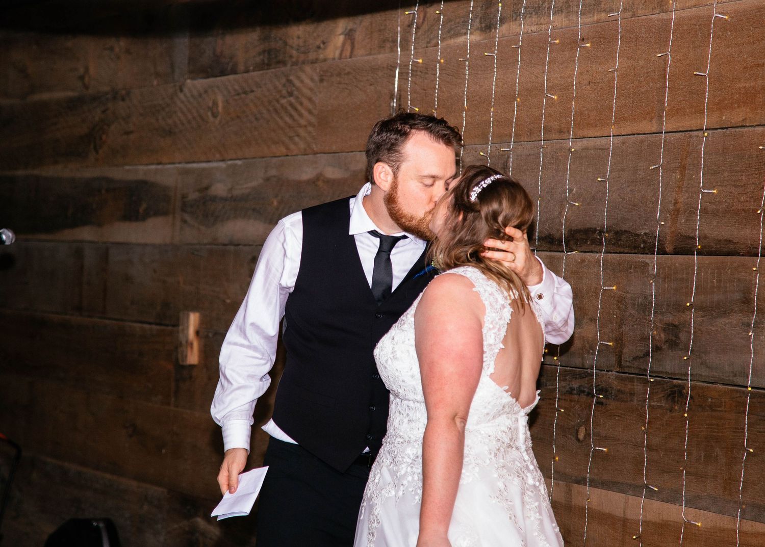 Bride and groom sharing a romantic kiss against a rustic wooden barn wall with string lights.