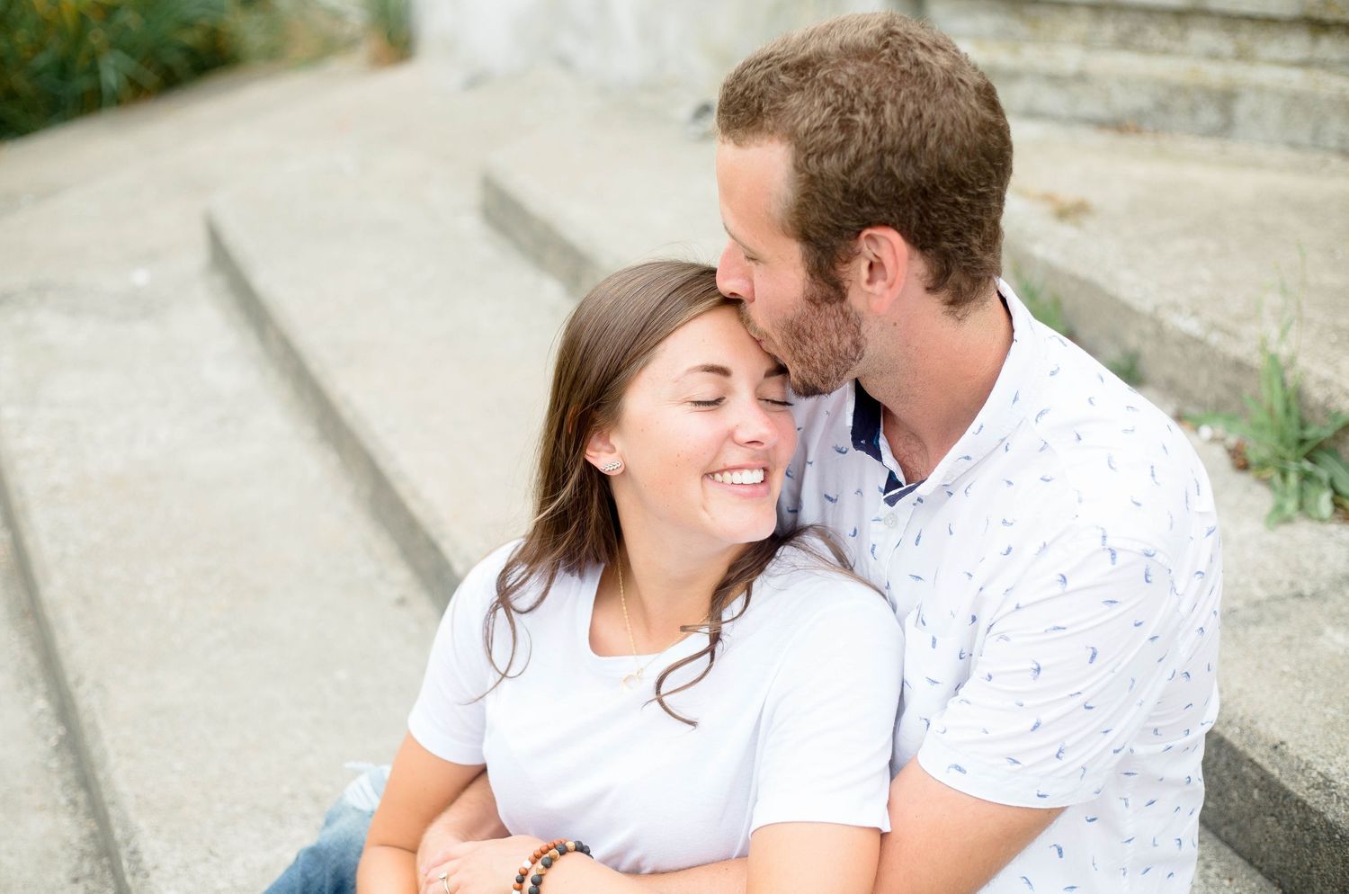 Man kissing smiling woman on the head while sitting on outdoor steps.