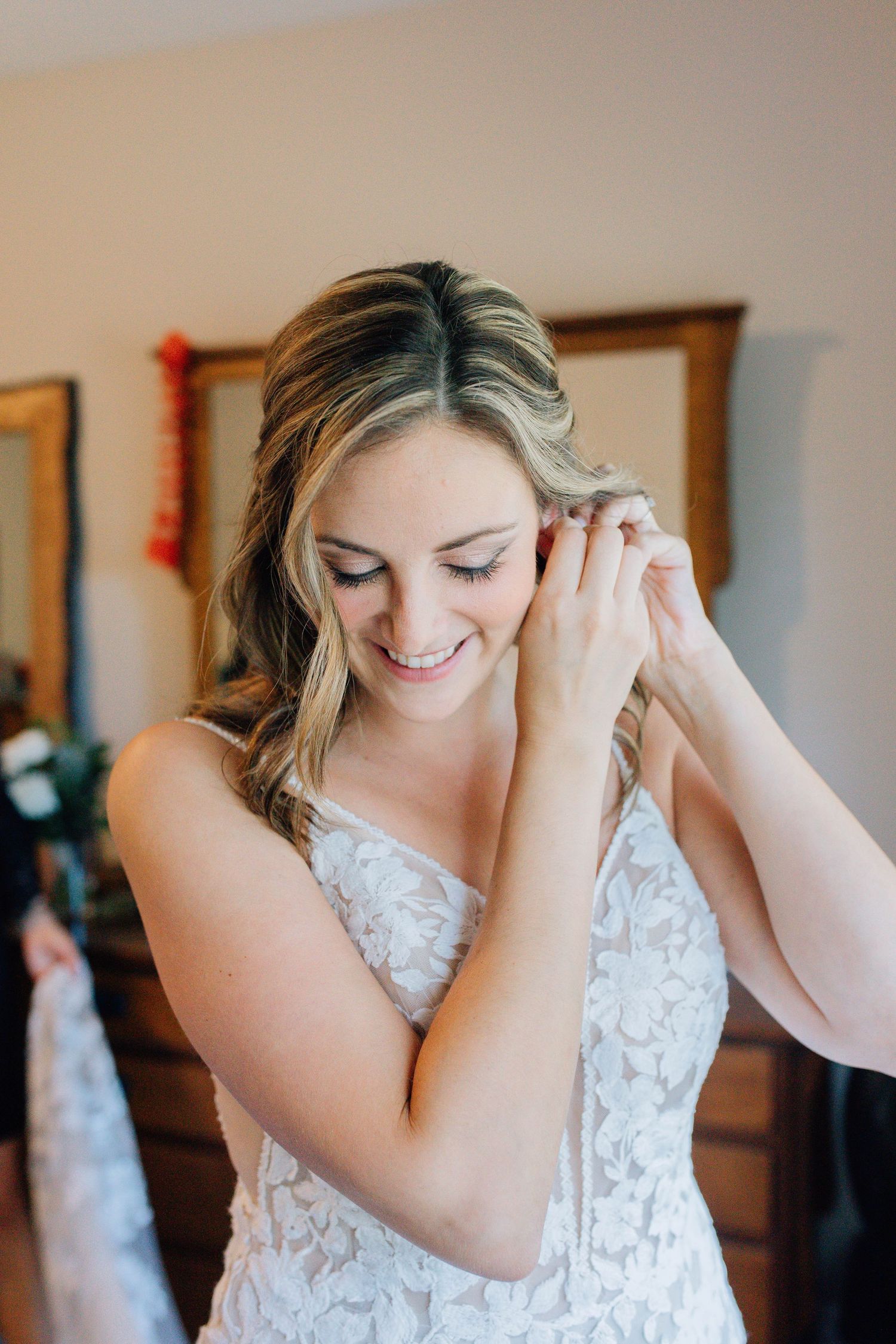Smiling bride in lace wedding dress putting on earrings in a warmly lit bridal suite.