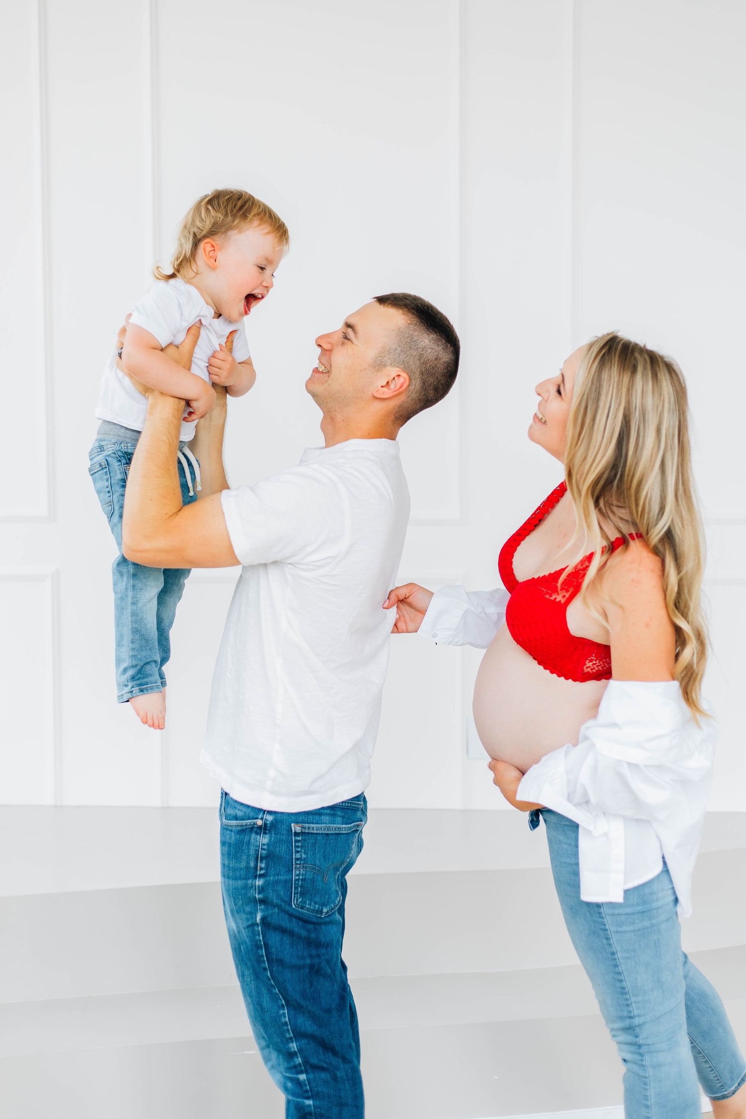 Happy family with pregnant mother, father lifting laughing toddler in white studio.
