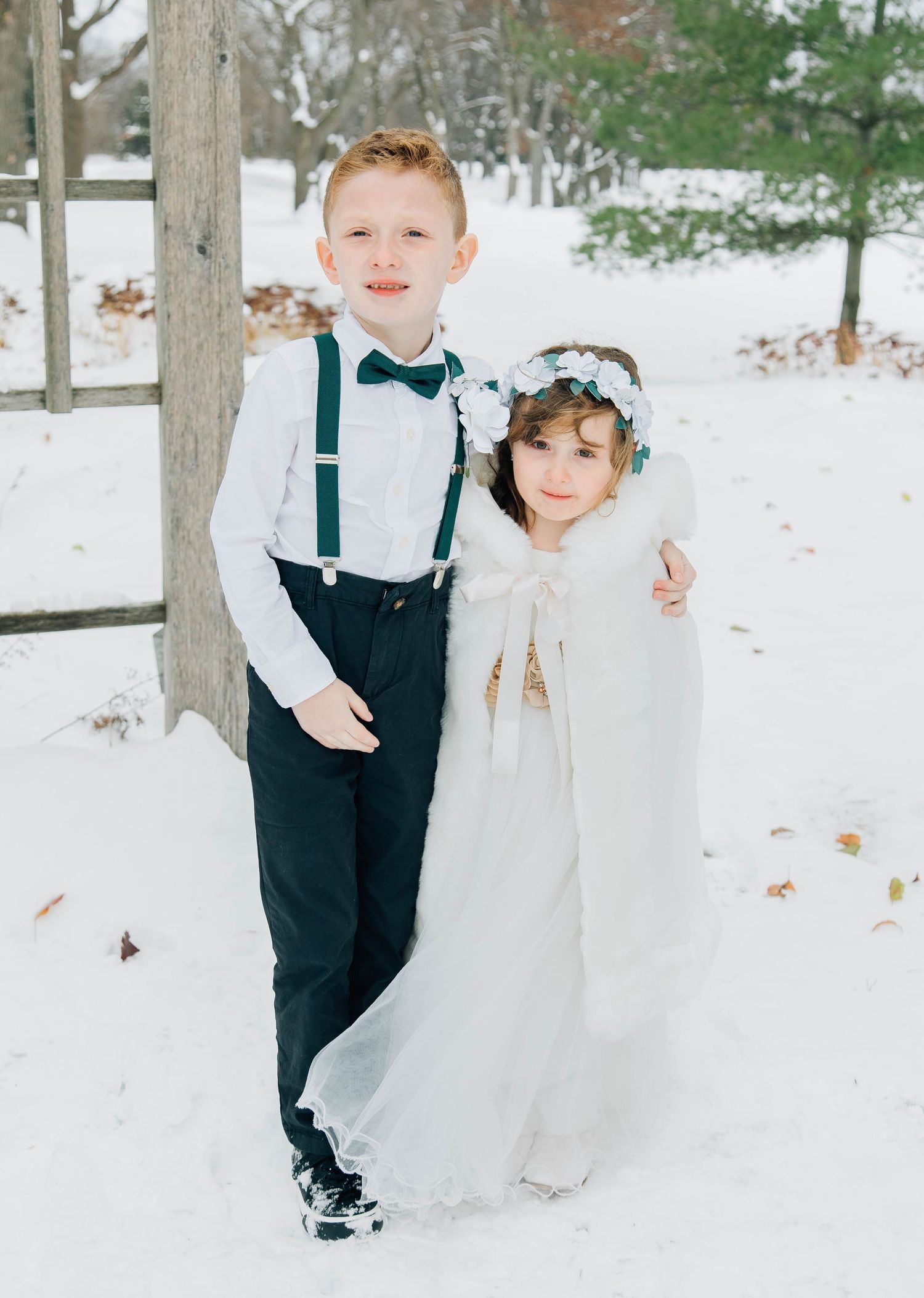 Ring bearer in suspenders and bow tie poses with flower girl in white cape and floral crown in snowy outdoor setting.