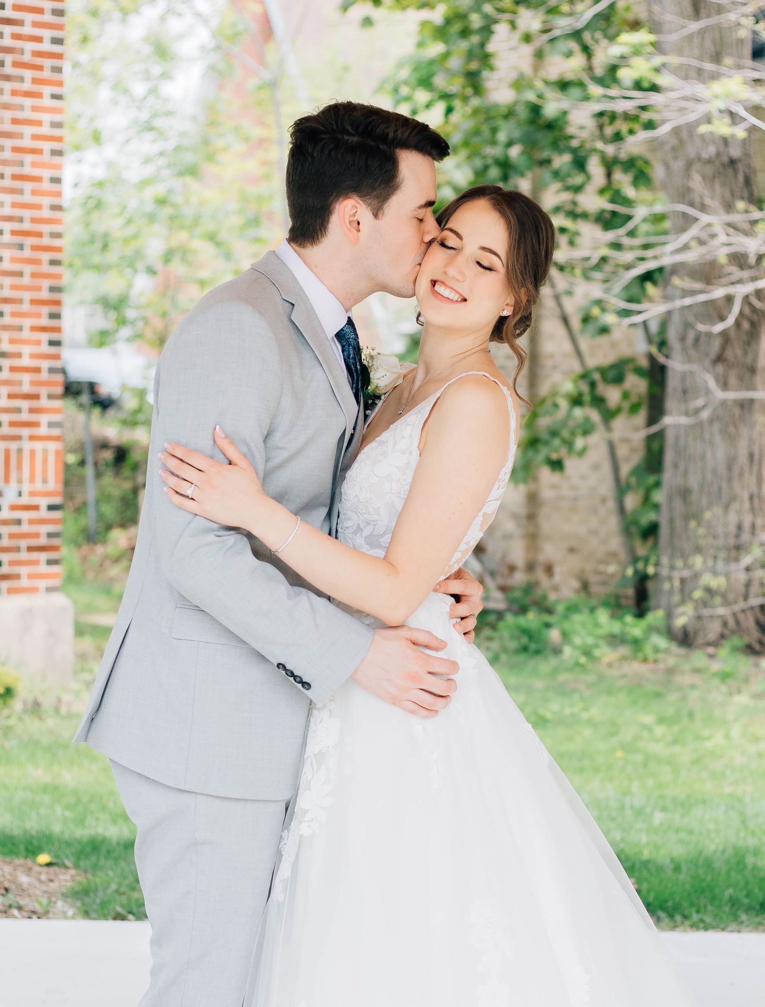 Groom in gray suit kisses smiling bride in white lace gown outdoors near brick building.
