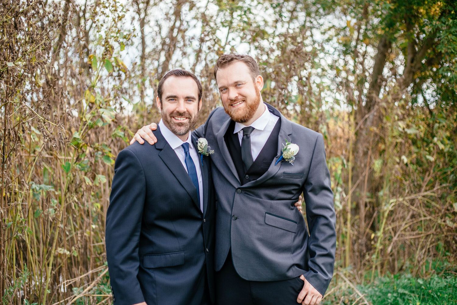 Two men in suits smiling together outdoors at wedding, one wearing boutonniere among autumn shrubs.