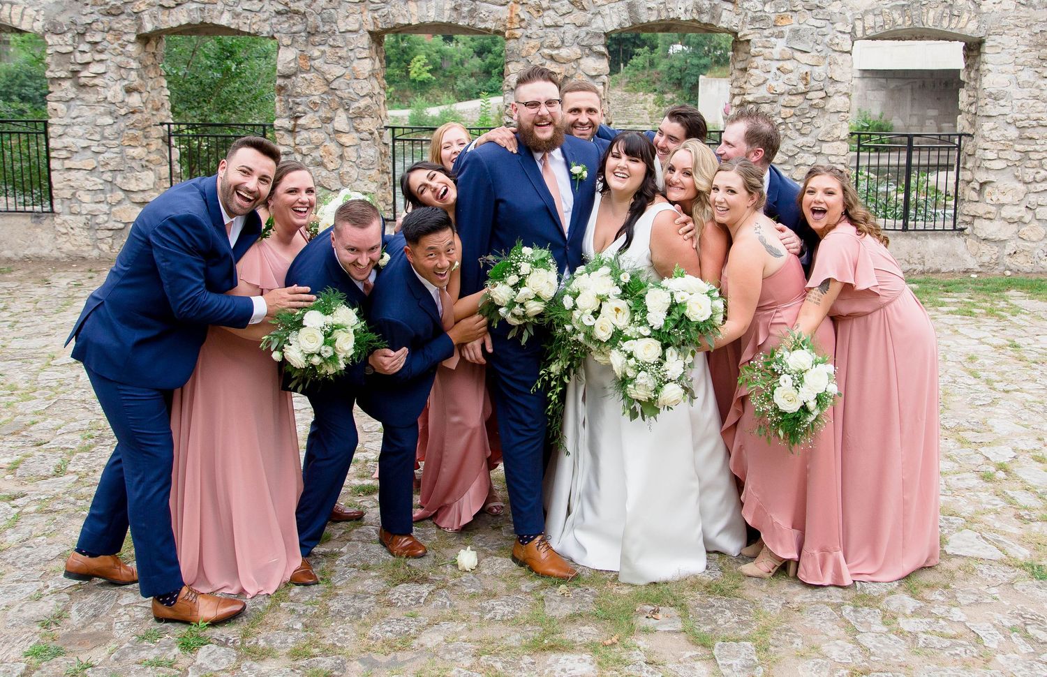 A joyful wedding party in navy suits and dusty rose dresses poses playfully in front of stone ruins.