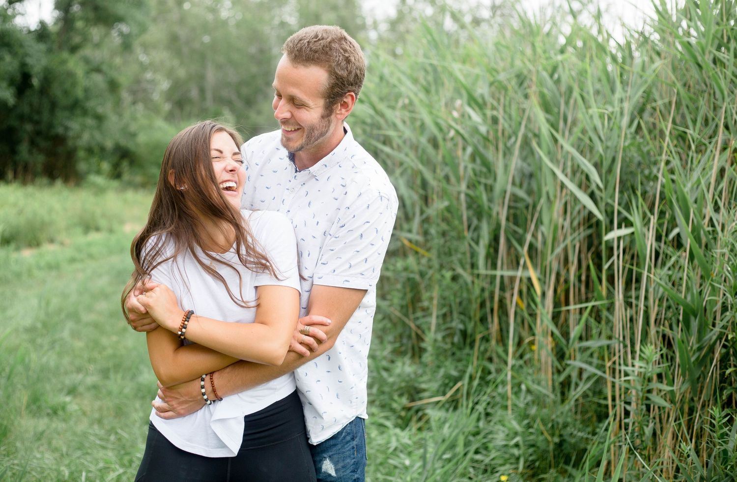 Couple embracing and laughing together near tall green reeds outdoors.