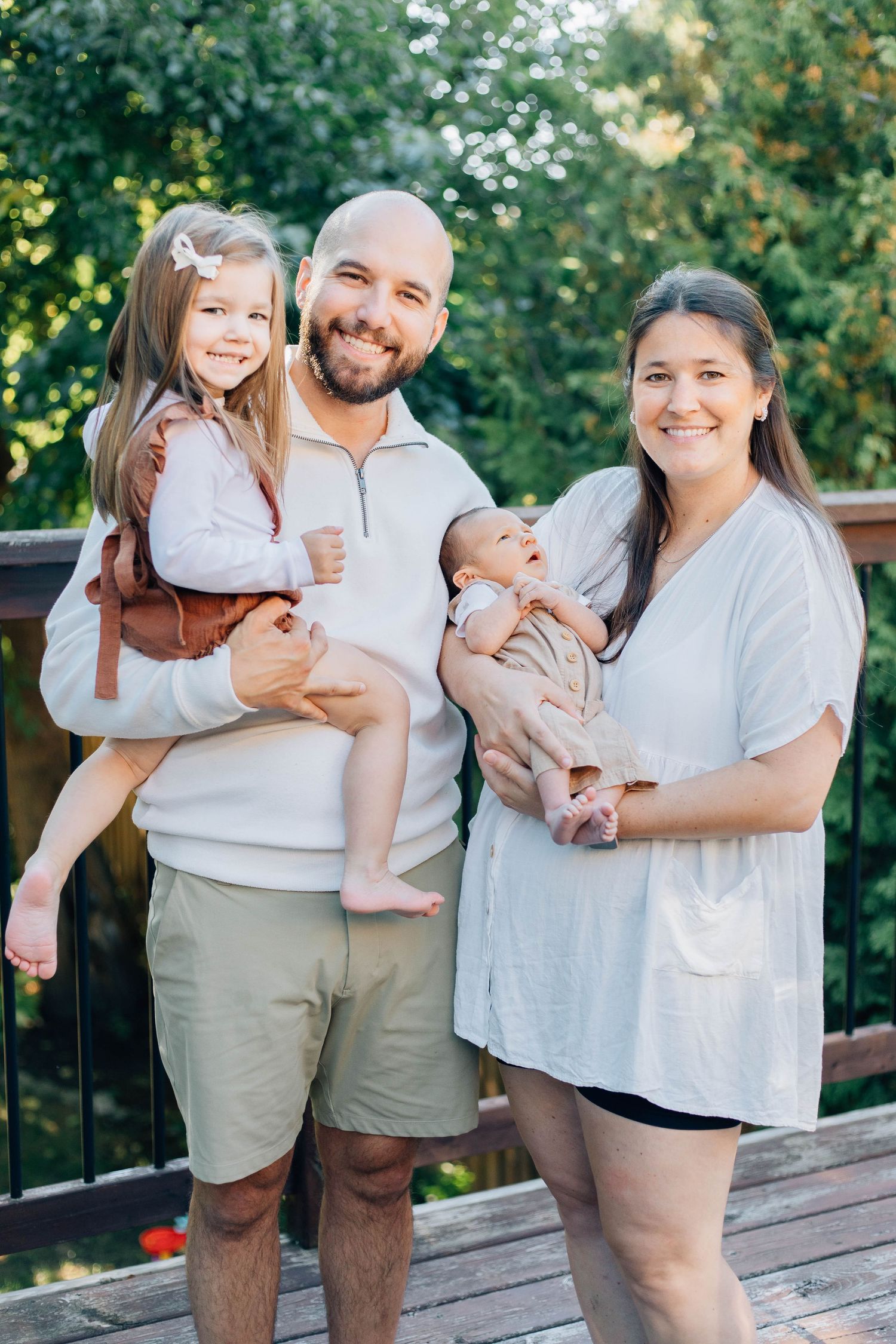 Happy family of four with toddler girl and newborn baby posing on wooden deck.