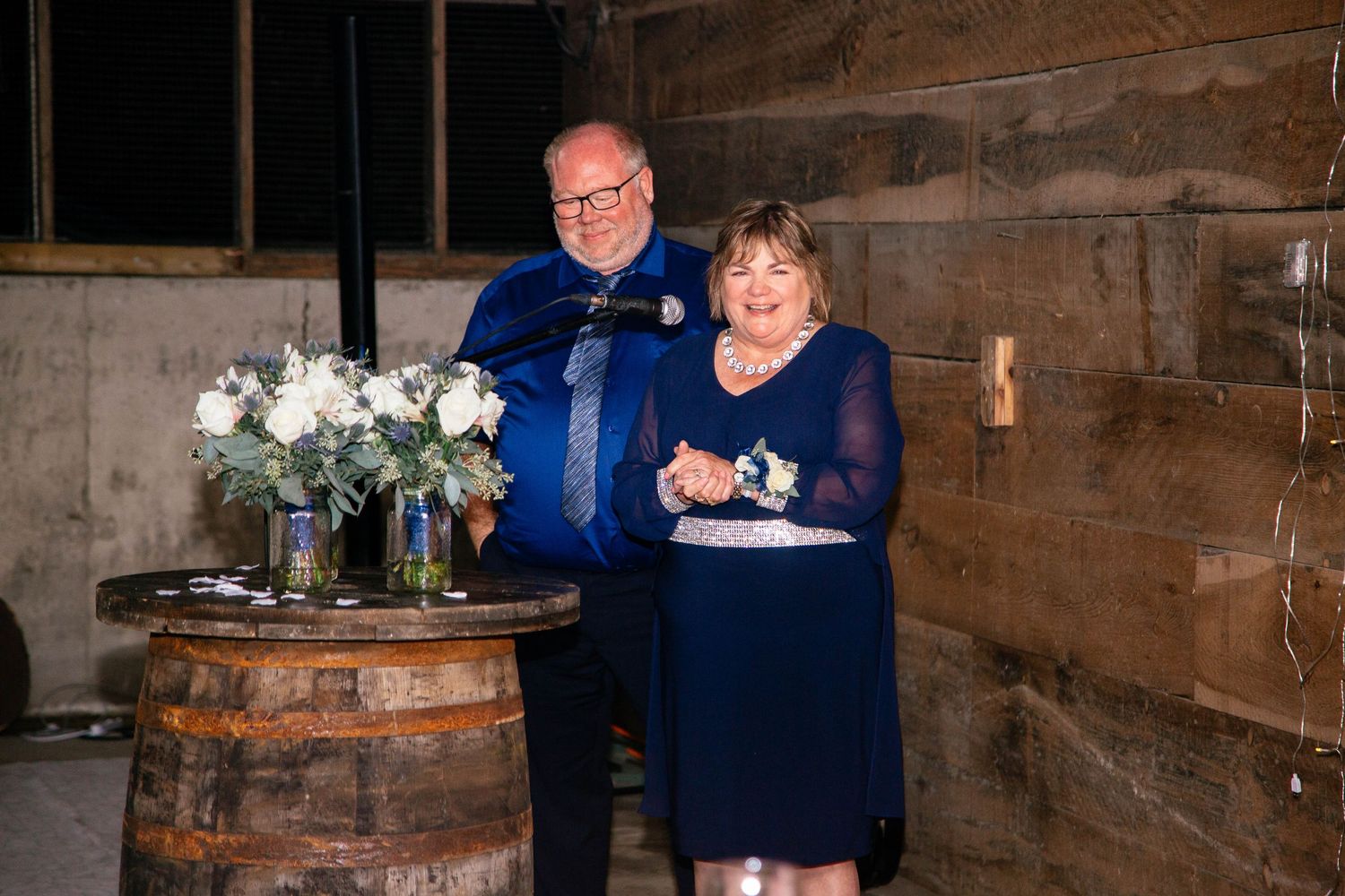 Couple in navy blue formal attire smiling together at rustic barn wedding reception barrel podium.