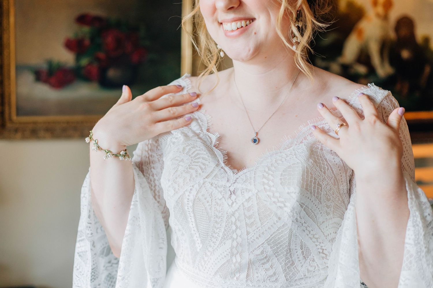 A smiling bride in a lace gown with bell sleeves touches her shoulders, showcasing her necklace and bracelet.