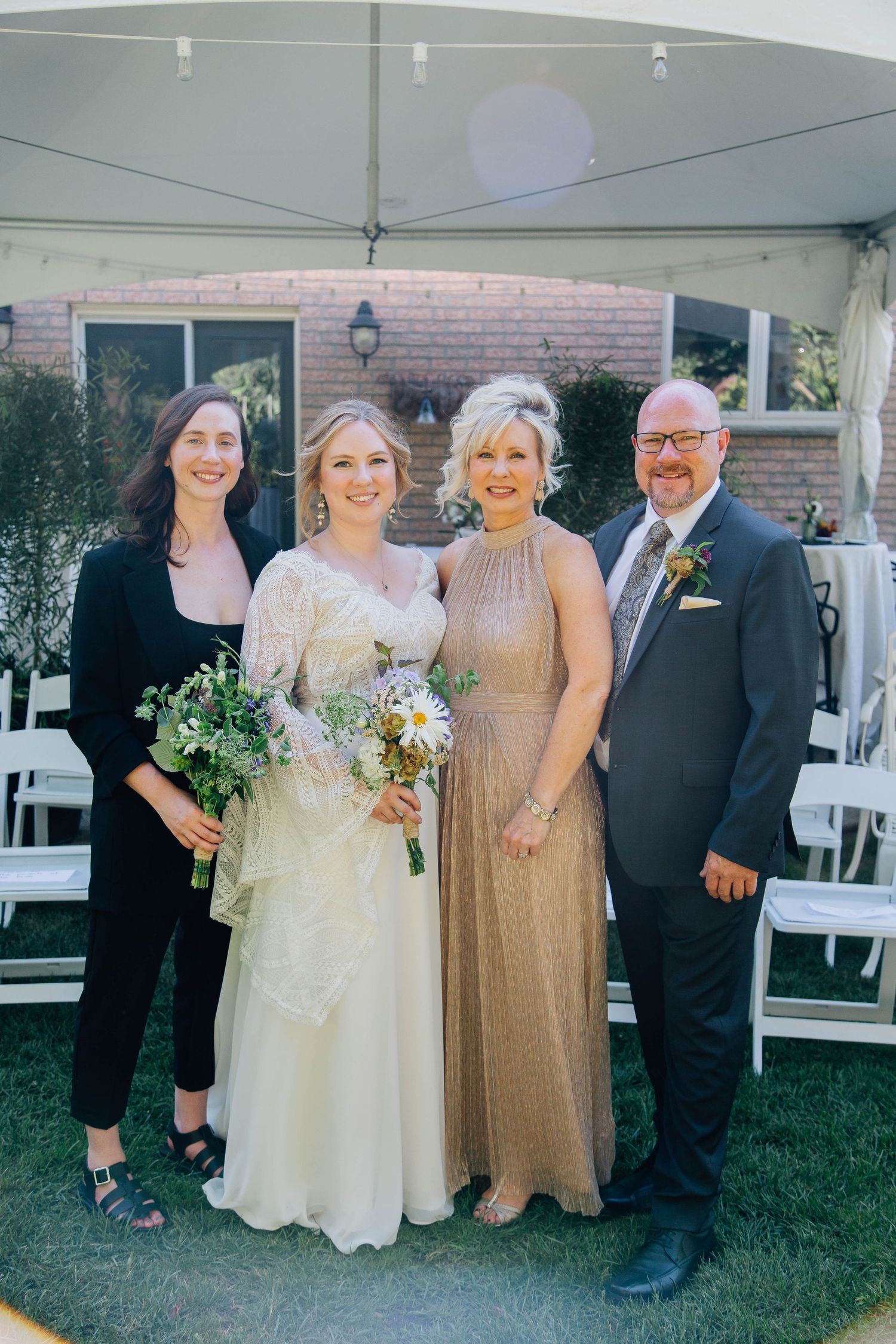 Bride in white lace gown poses with three family members including mother in gold dress and father in suit at backyard wedding.