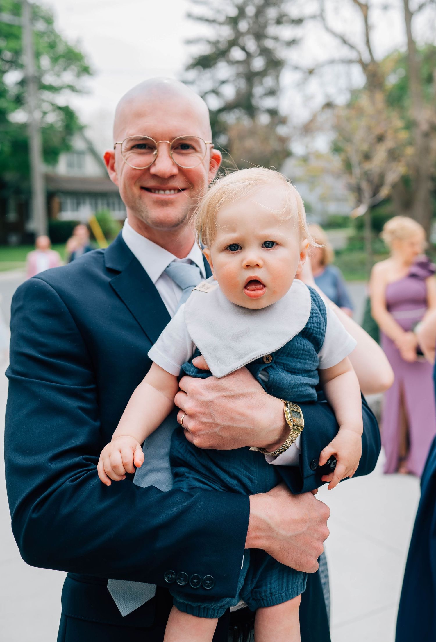 Smiling bald man in navy suit holding an adorable baby in denim overalls.