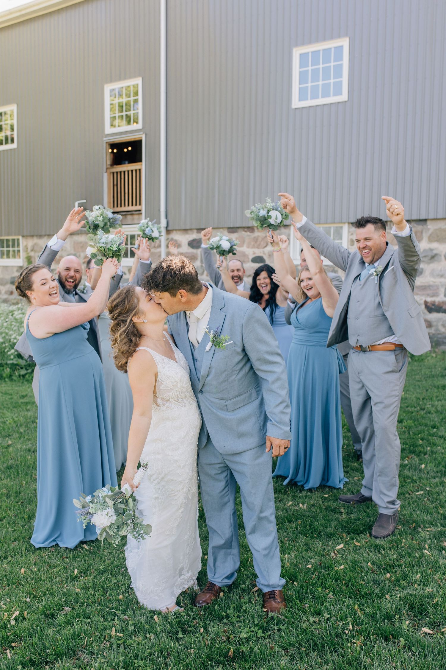 Bride and groom kiss surrounded by cheering wedding party in blue dresses and gray suits outside a barn venue.