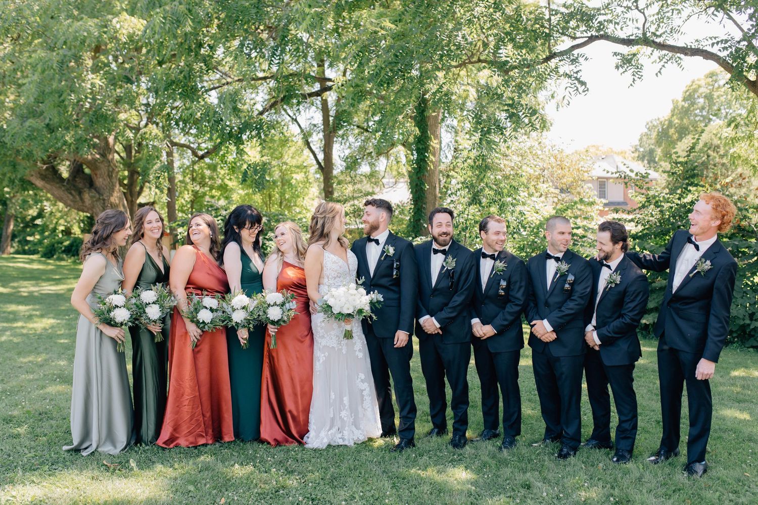 Wedding party in mismatched bridesmaid gowns and black tuxedos posing outdoors under trees.