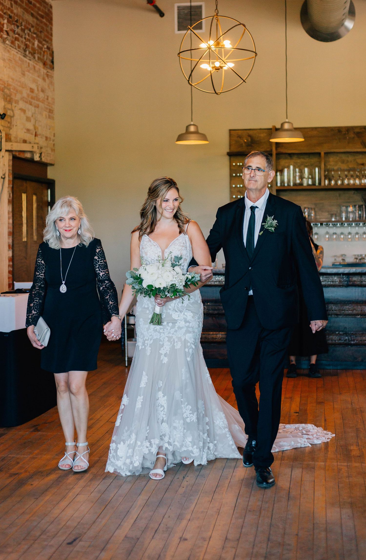 Bride in lace mermaid gown walks down aisle escorted by parents in rustic industrial wedding venue.