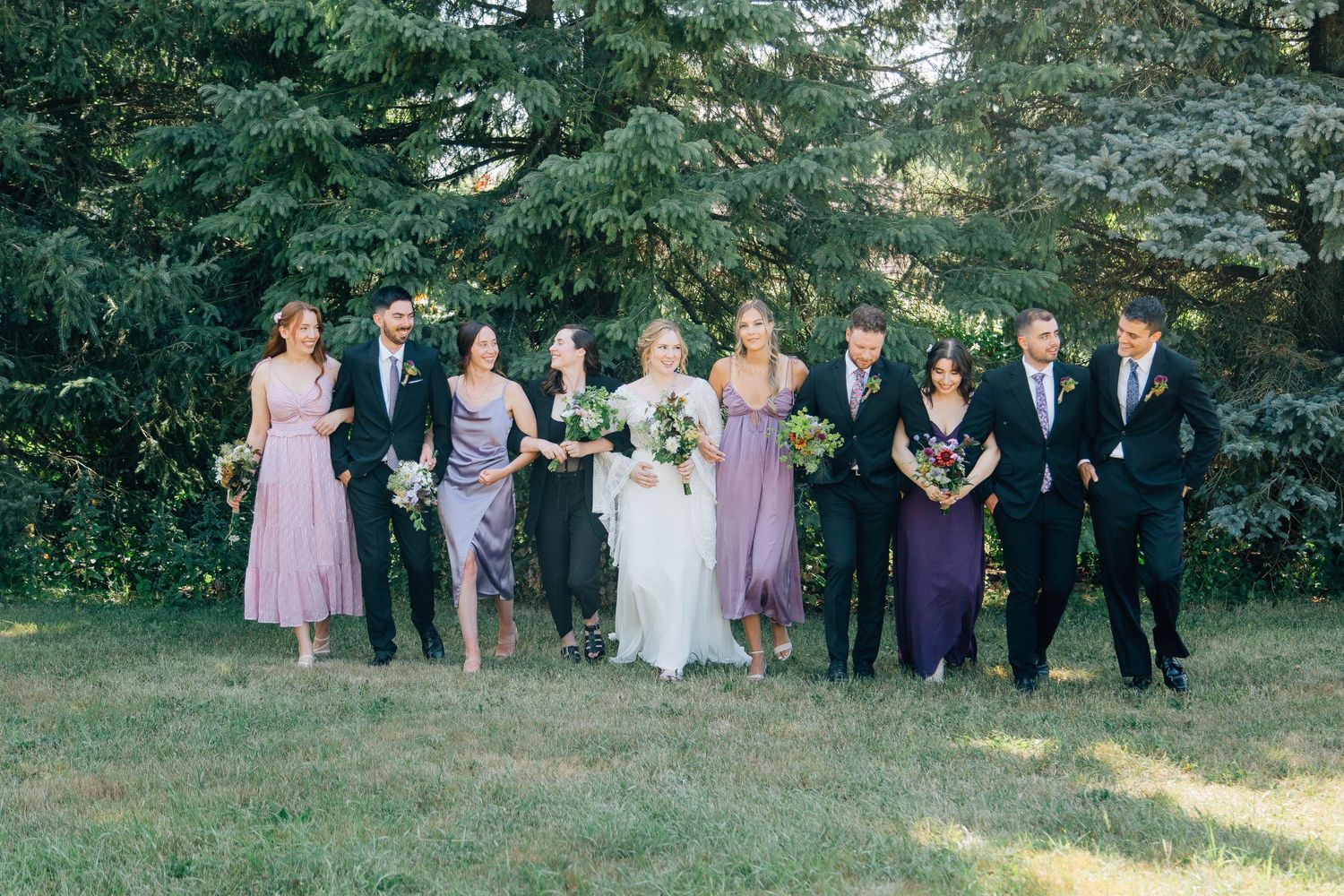 Wedding party in purple dresses and black suits walking together on green lawn with evergreen backdrop.