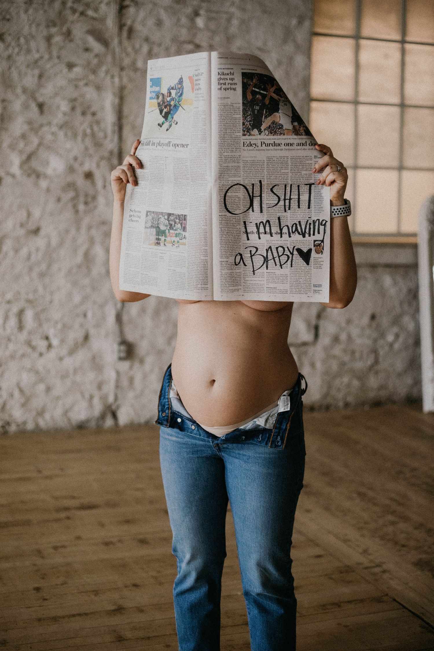 Pregnant woman holding a newspaper with handwritten text reading oh shit Im having a baby over her face.