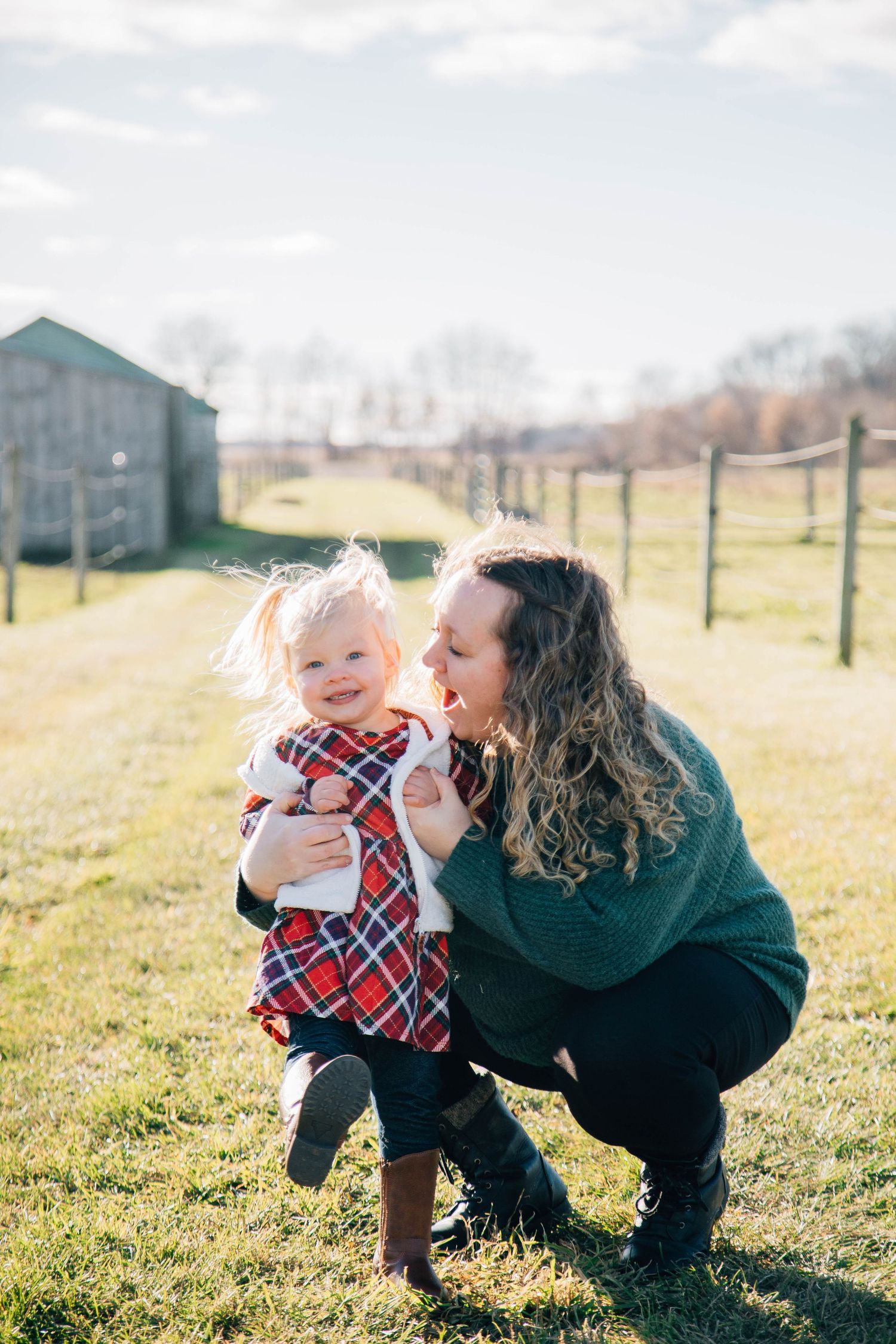 Mother crouching down smiling with her toddler daughter in a plaid dress on a farm field.