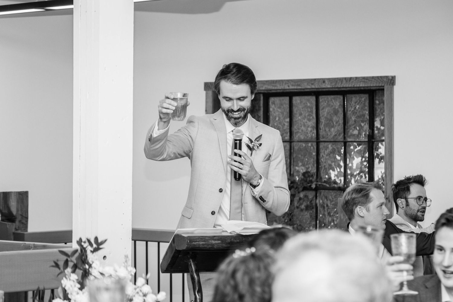 Man in suit smiling and gesturing while giving a speech at a wedding reception.