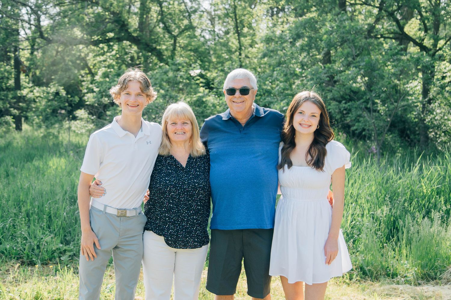 Family of four, two adults and two teenagers, smiling together in a green outdoor setting on a sunny day.