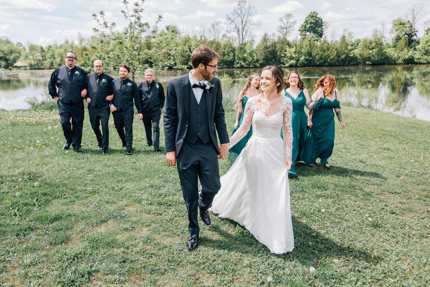 Bride and groom walking hand in hand on grass while wedding party follows behind near a lake.