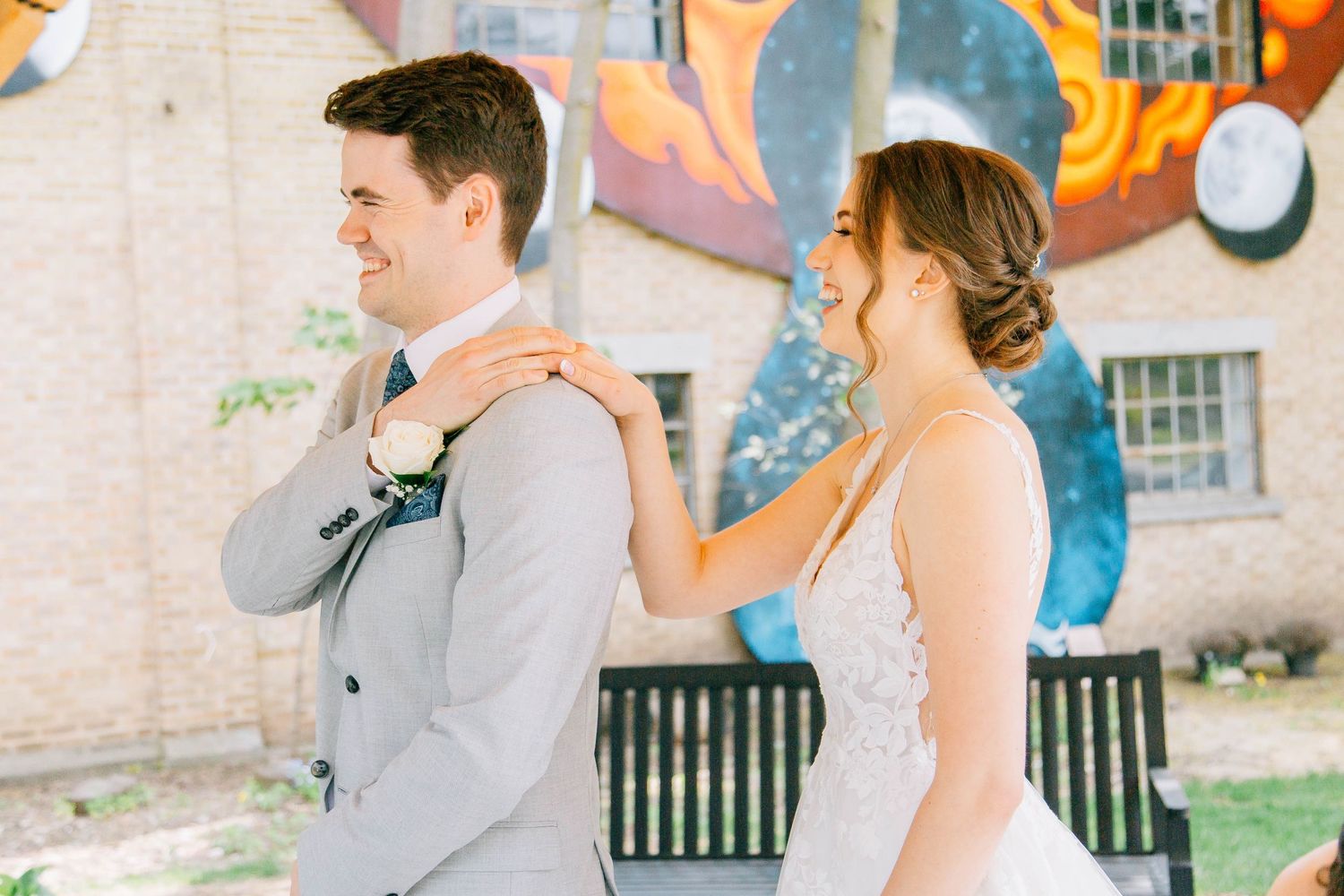 Bride in lace gown places hands on grooms shoulders during first look at colorful mural backdrop.
