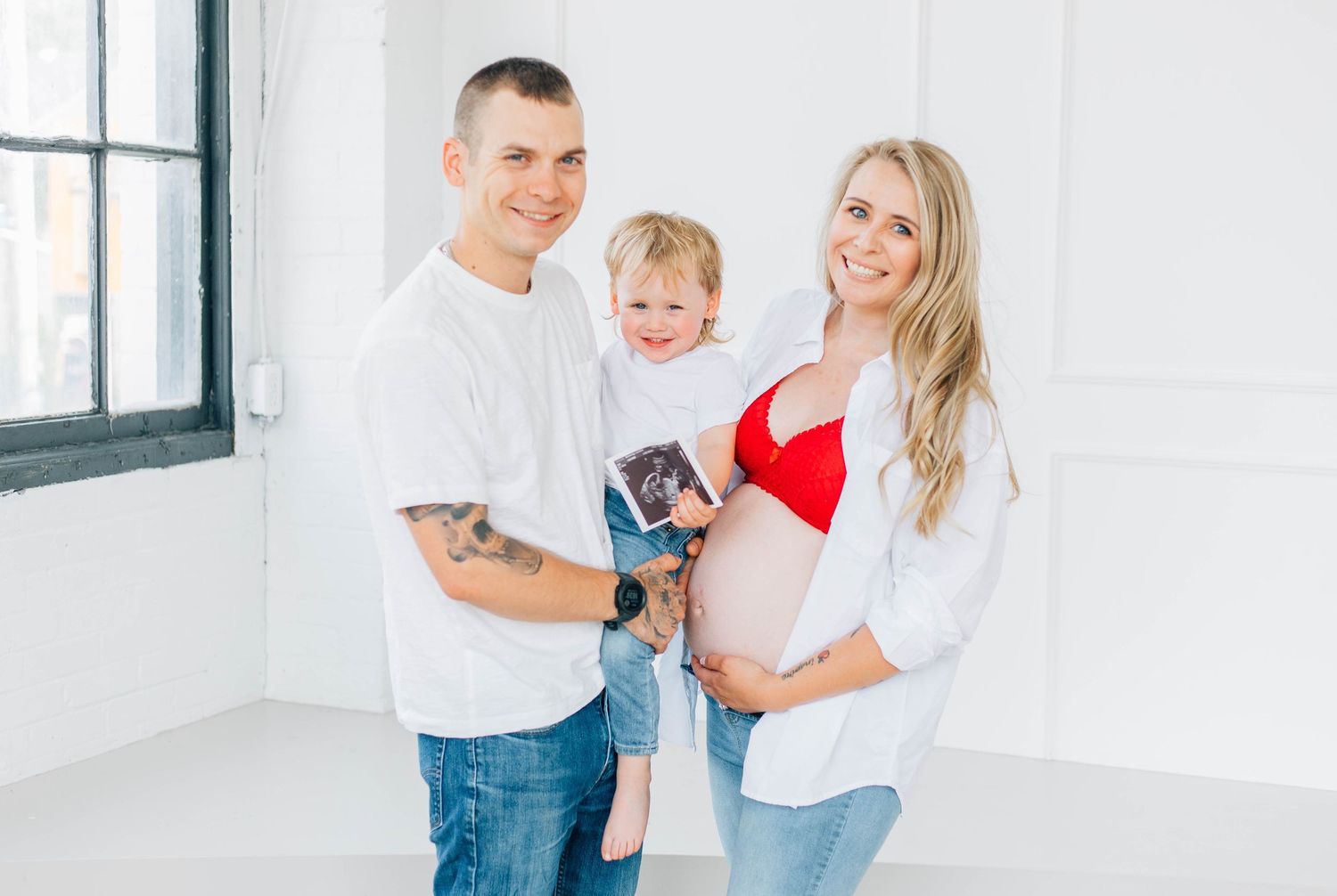 Happy family maternity photo with pregnant mom in red bralette holding ultrasound with toddler and tattooed dad.
