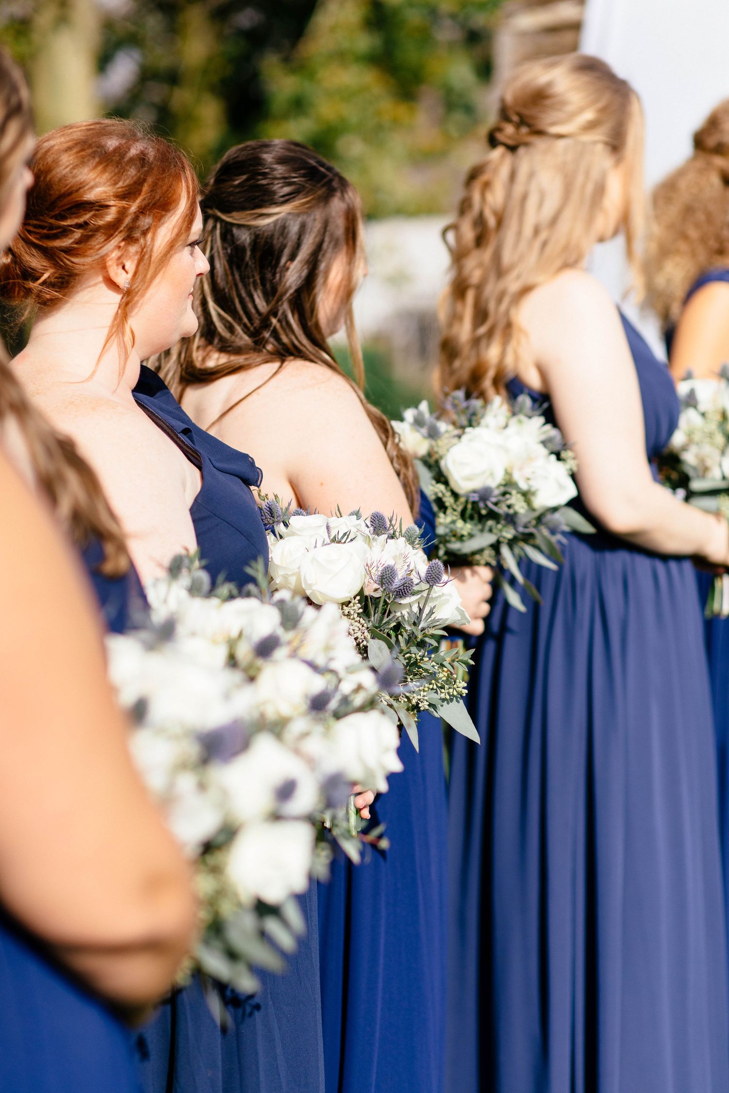 Bridesmaids in navy blue gowns hold white and green bouquets at an outdoor wedding.