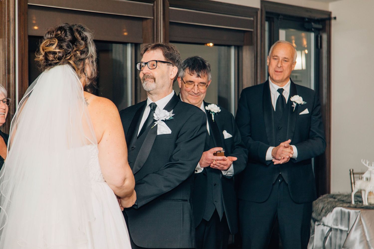 Groomsmen in black tuxedos smile and clap during indoor wedding ceremony beside large windows.