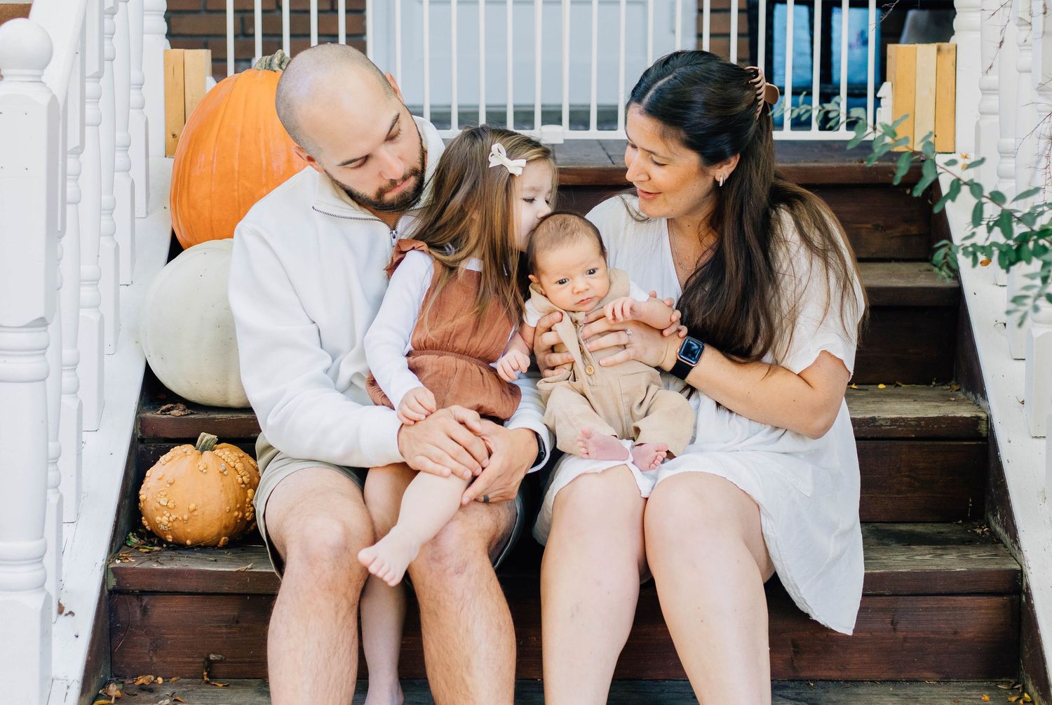 Family of four sitting on fall porch steps with pumpkins, parents smiling at newborn baby while toddler daughter sits nearby.