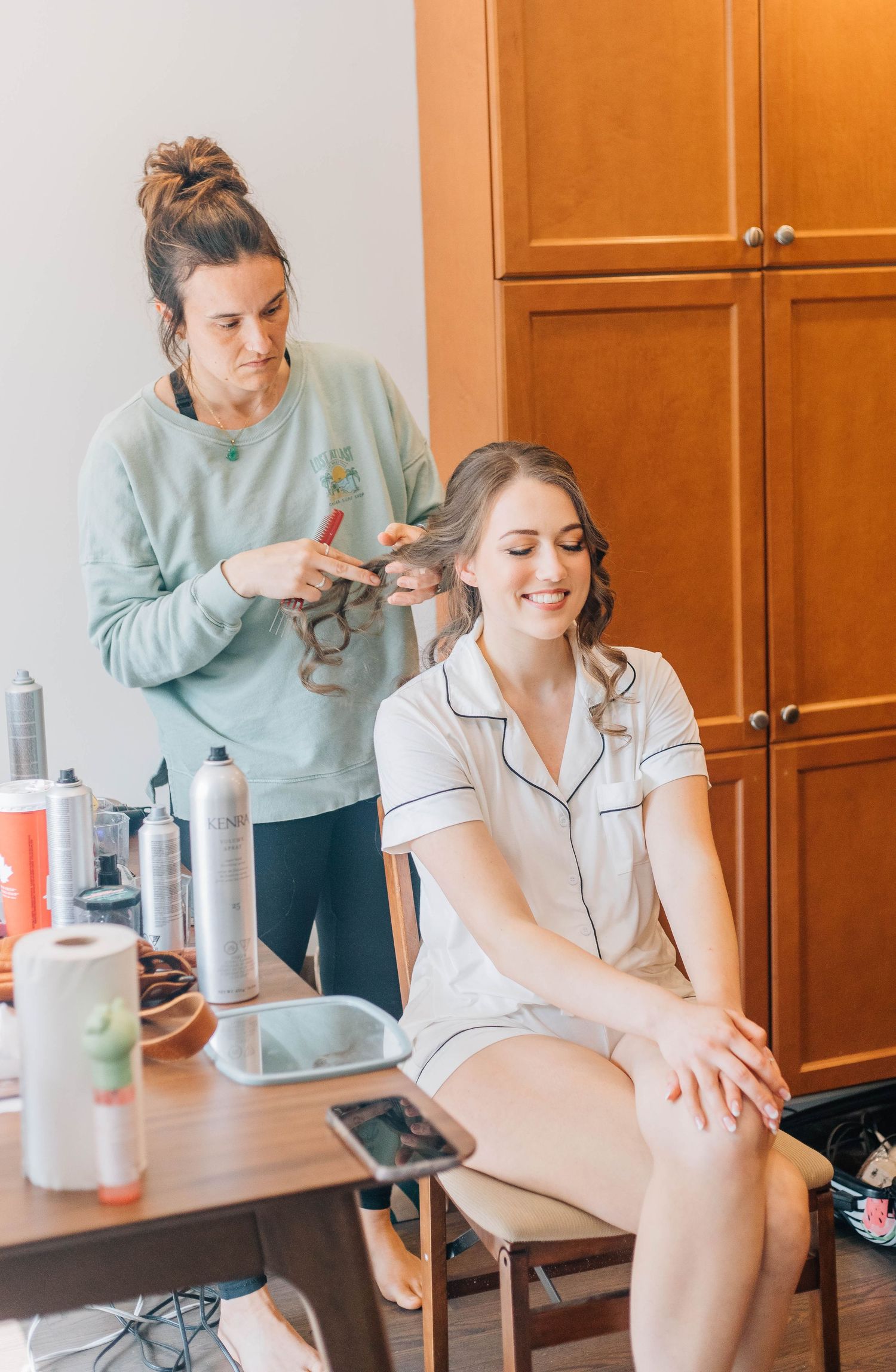 Hair stylist styling bride's hair while she smiles in white pajamas before wedding.
