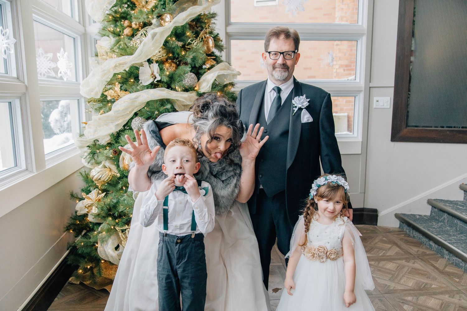 Bride and groom pose with young flower girl and ring bearer in front of decorated Christmas tree at winter wedding.