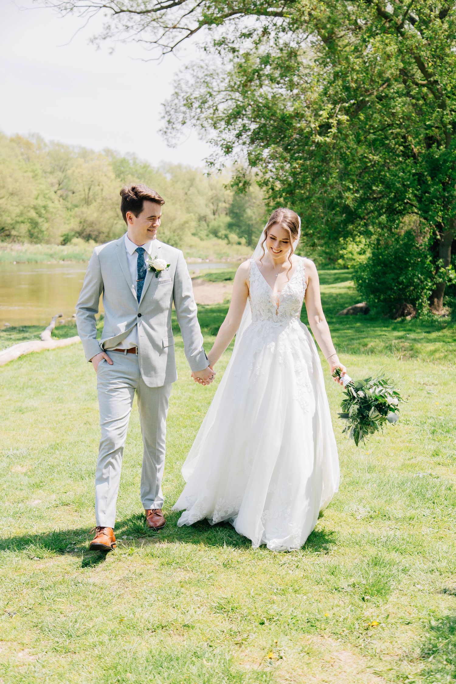Happy newlyweds walking hand in hand on green grass near a river, bride holding a lush bouquet.