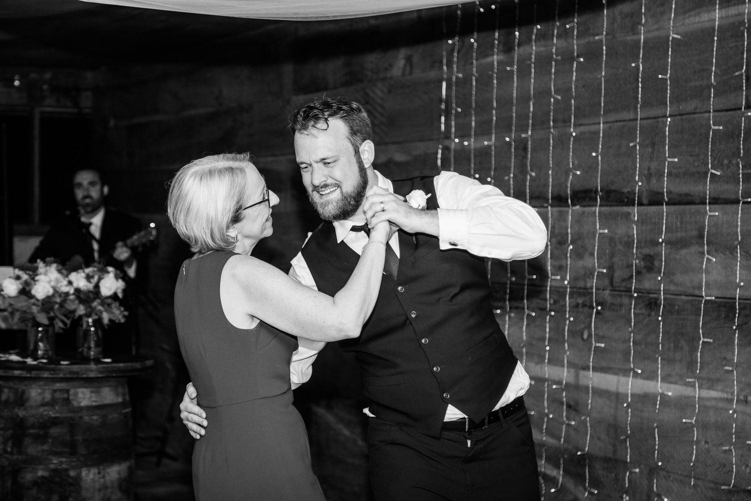 Black and white photo of groom sharing emotional mother-son dance at rustic barn wedding reception.
