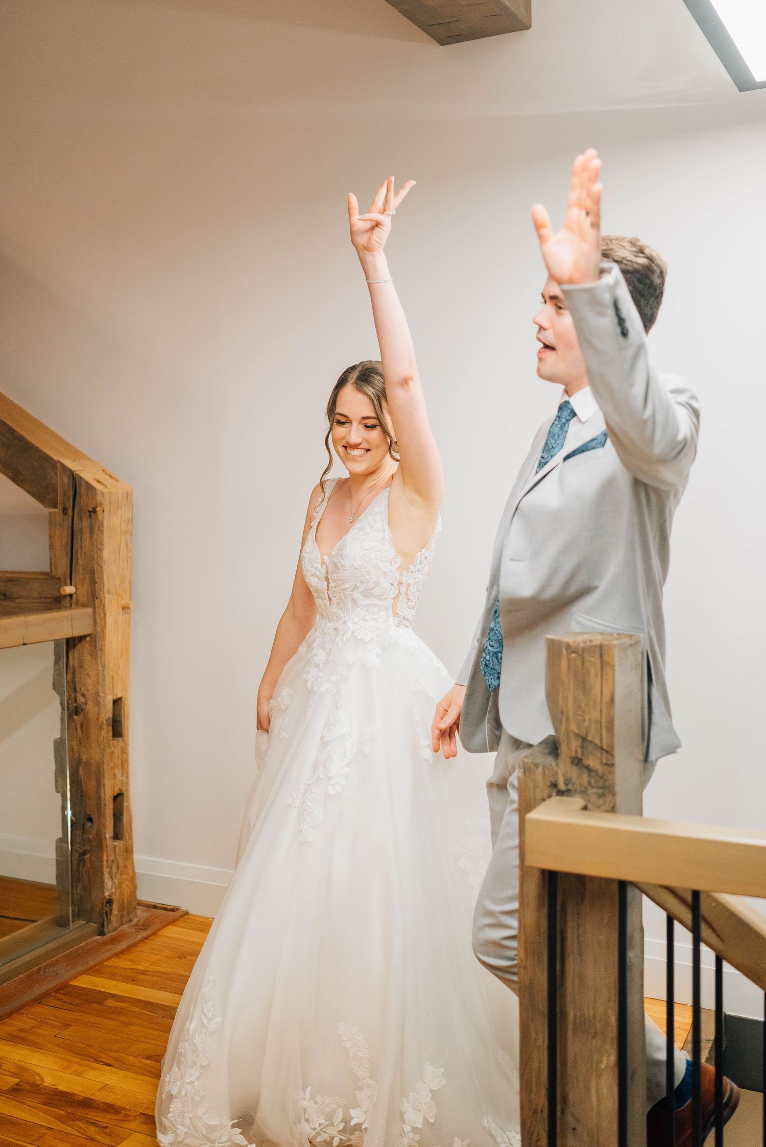 Excited bride in white lace gown and groomsman celebrating with raised hands at wedding reception.